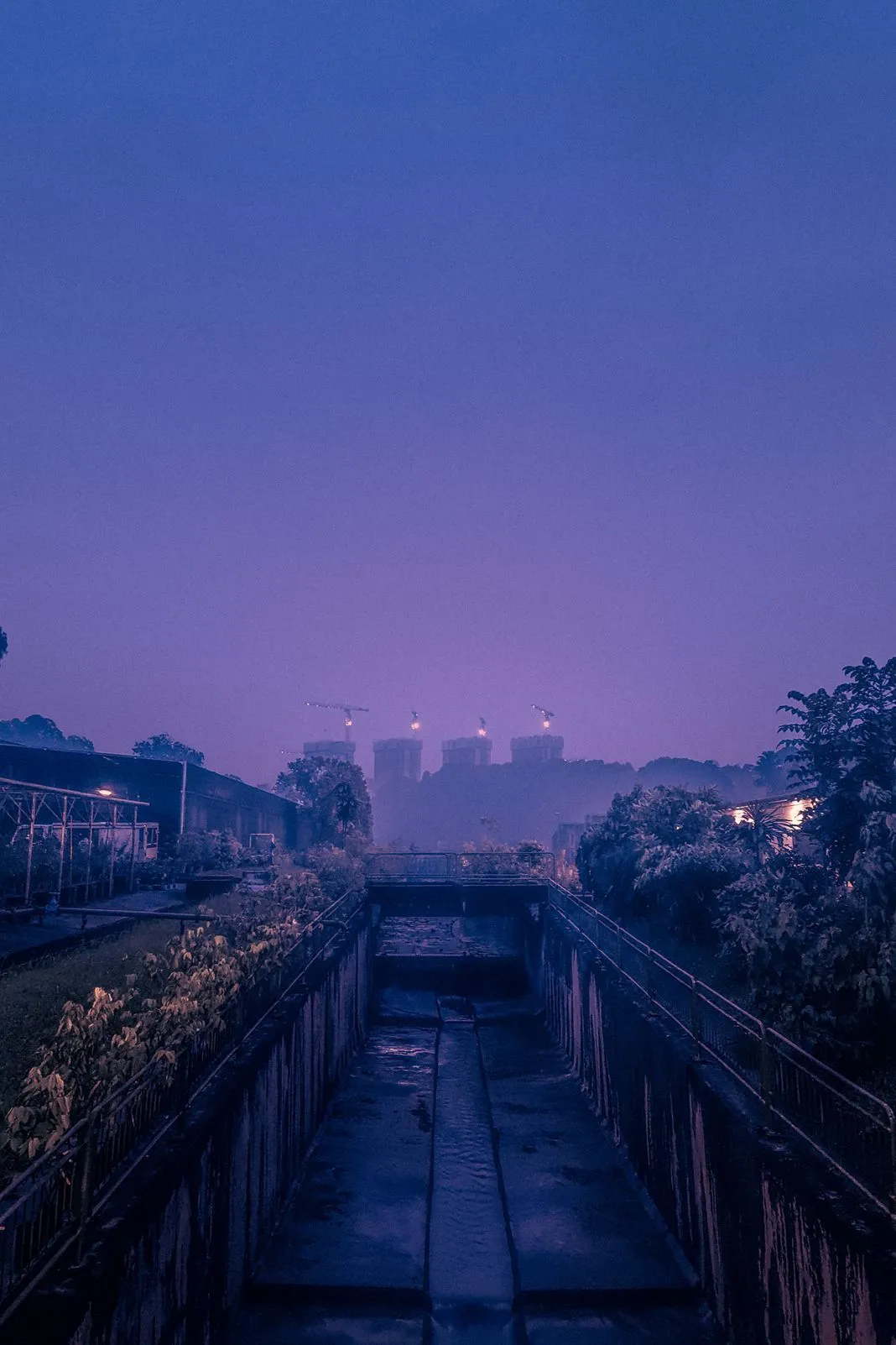 A concrete drainage canal leading toward buildings under construction at twilight