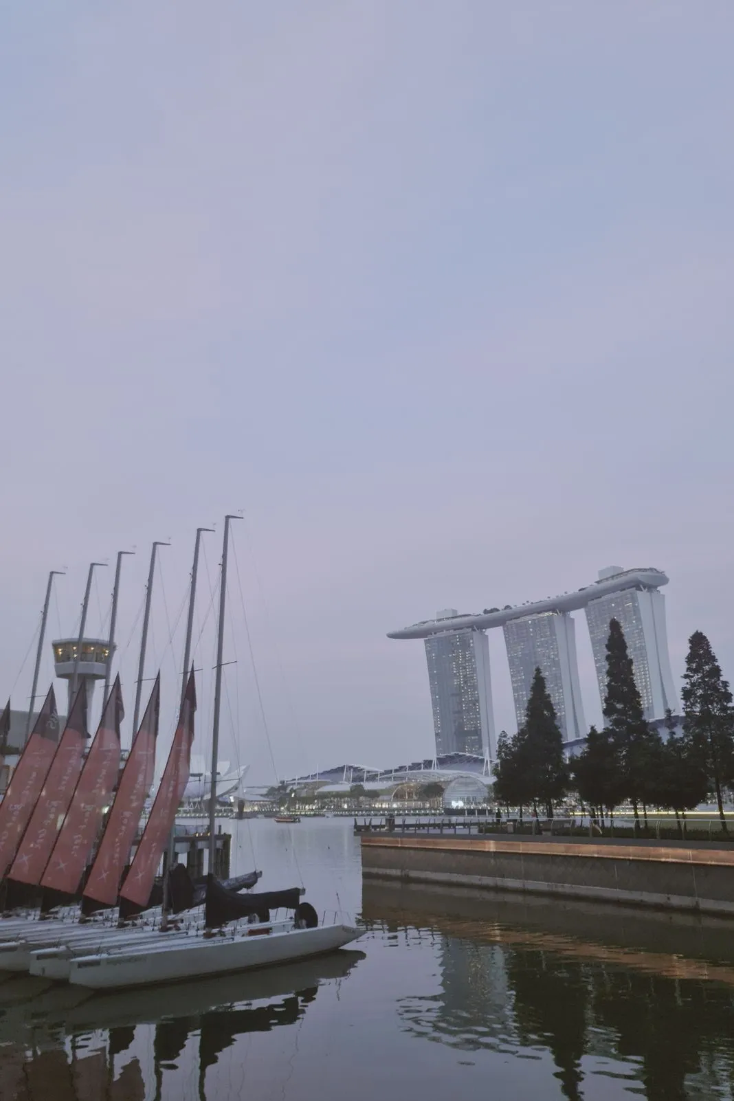 Red-flagged sailboats moored in a marina with Marina Bay Sands beyond