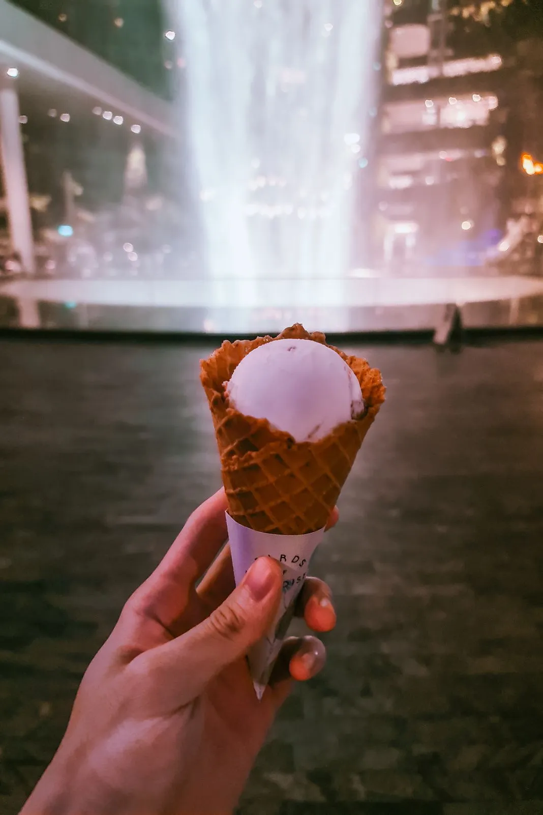 A hand holding a waffle cone ice cream before a lit indoor waterfall at night