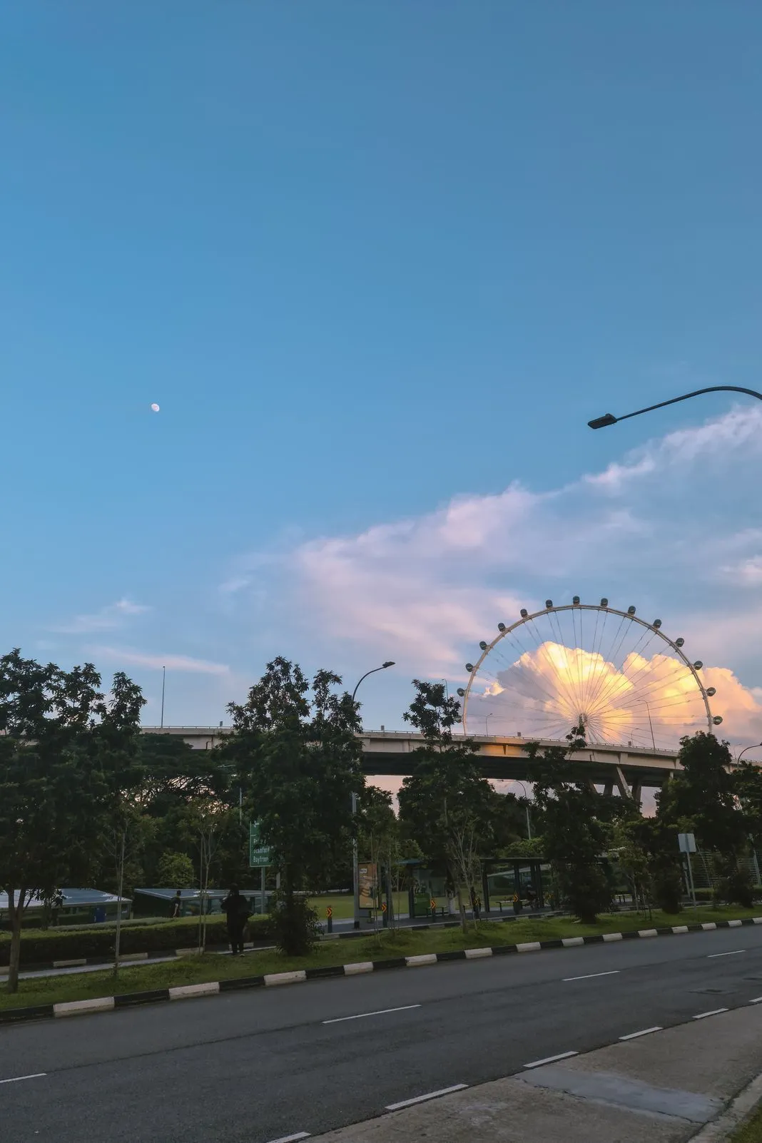 A ferris wheel rising behind clouds above an overpass, moon visible in daytime sky