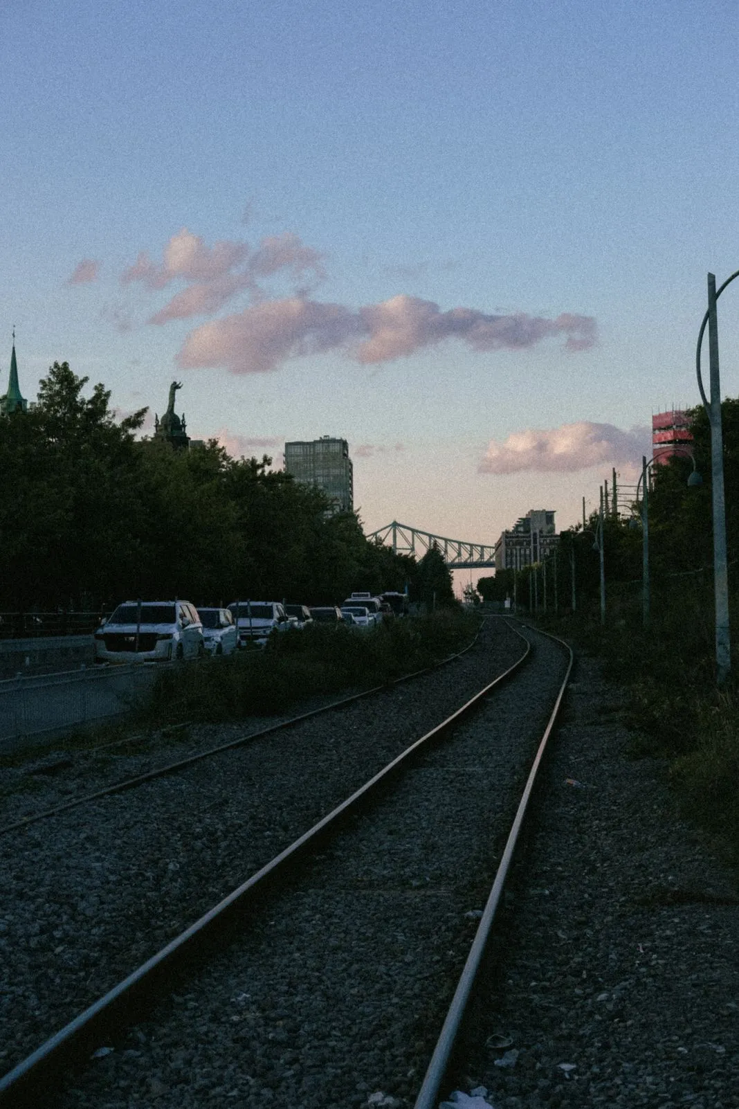 Railway tracks curving past parked cars toward a suspension bridge at dusk