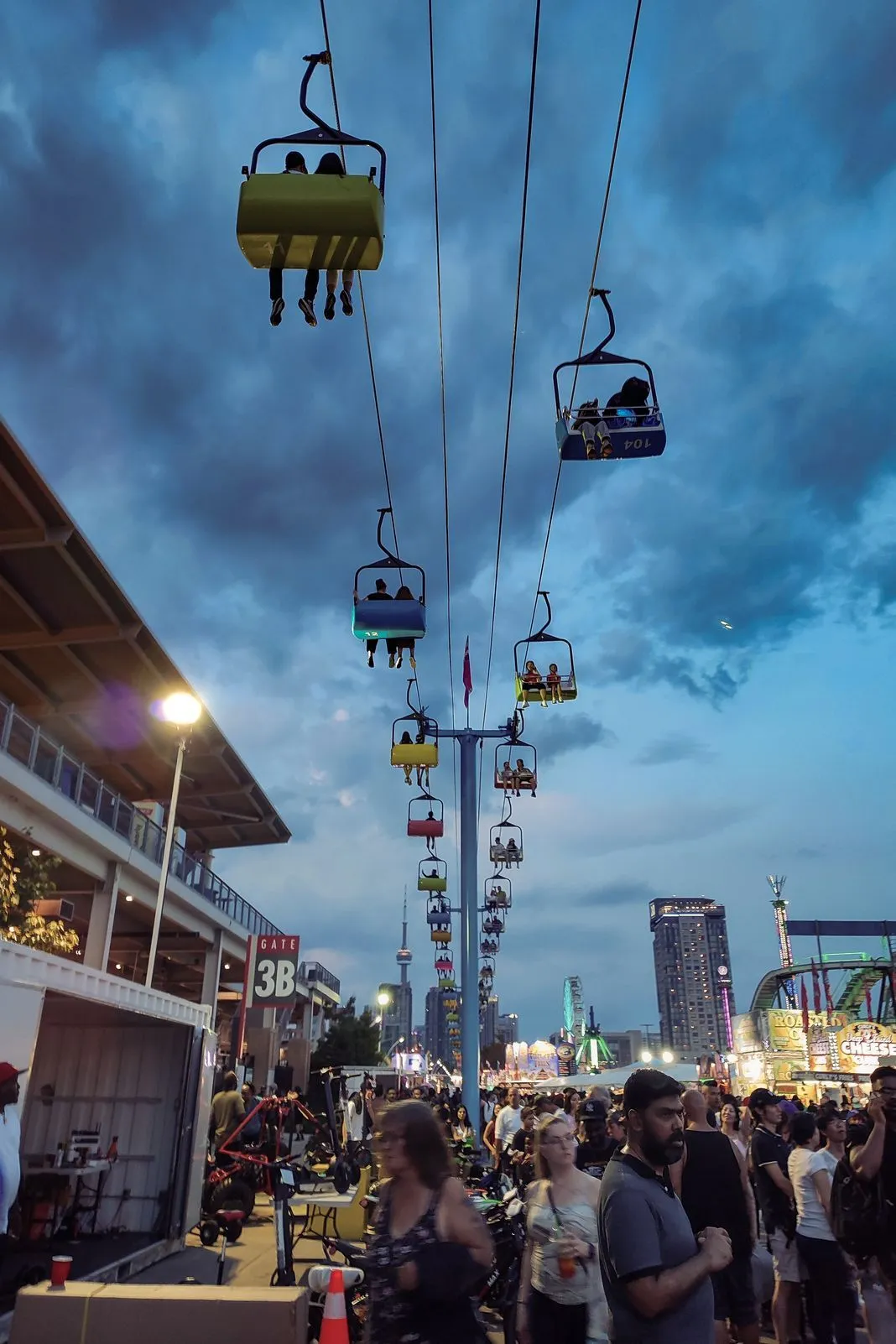Chairlift gondolas swinging above a crowded fairground at dusk with the skyline behind