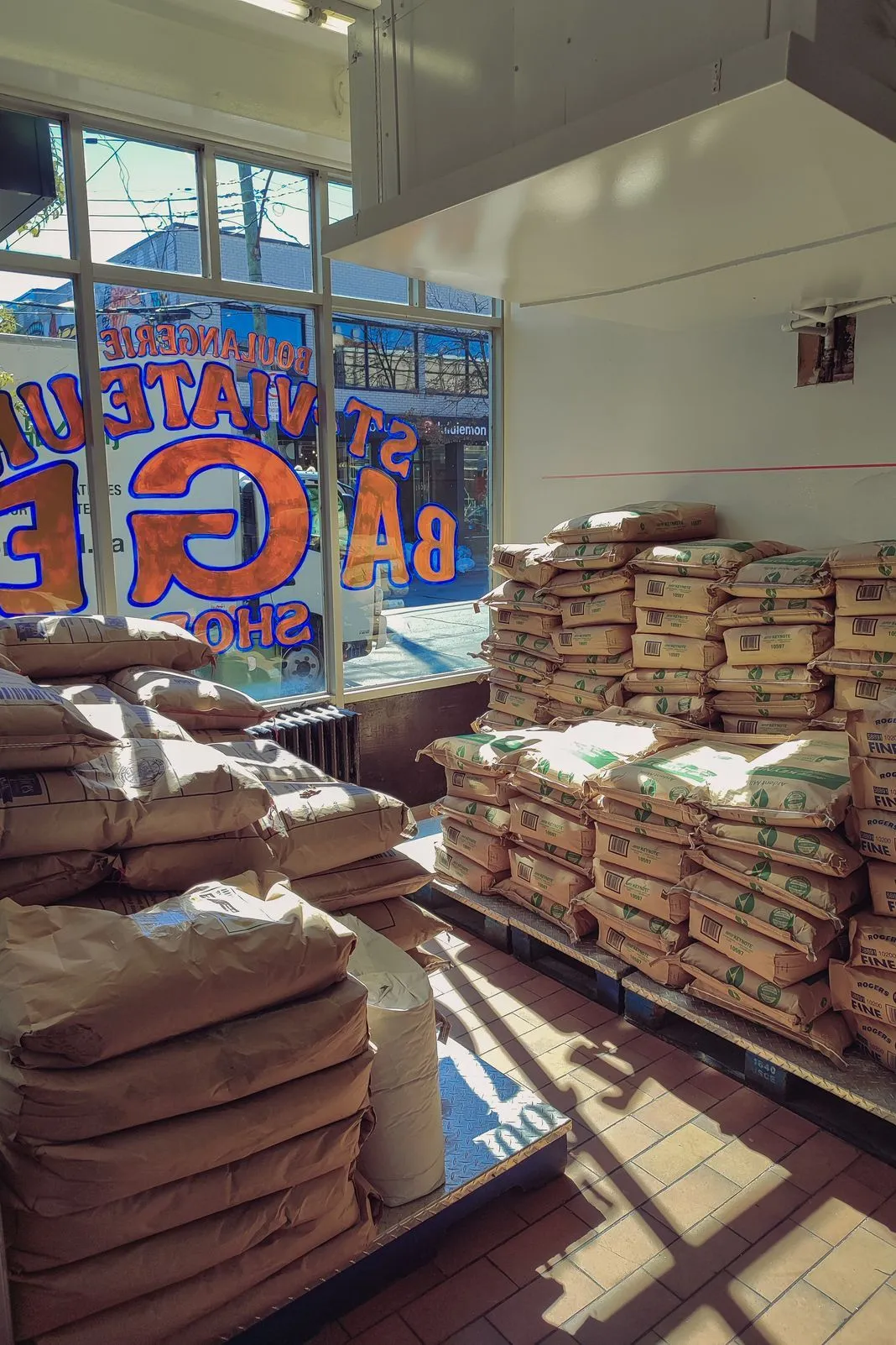 Stacked flour sacks inside a bakery supply shop with a neon sign in the window