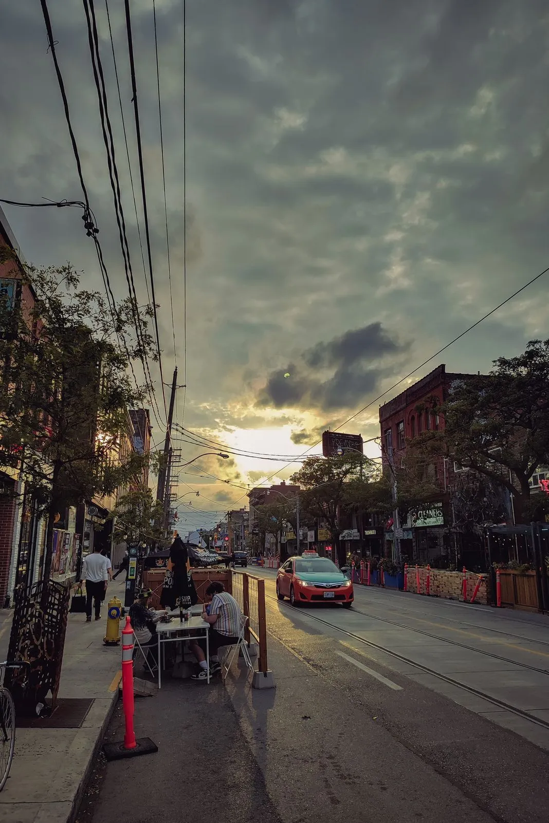 Outdoor patio diners and a taxi on a city street lit by a cloudy sunset
