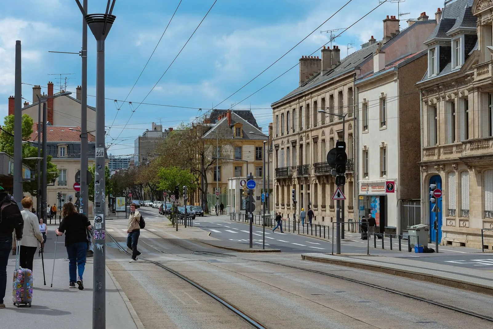 Pedestrians and a rolling suitcase on a street with tram tracks and ornate stone buildings