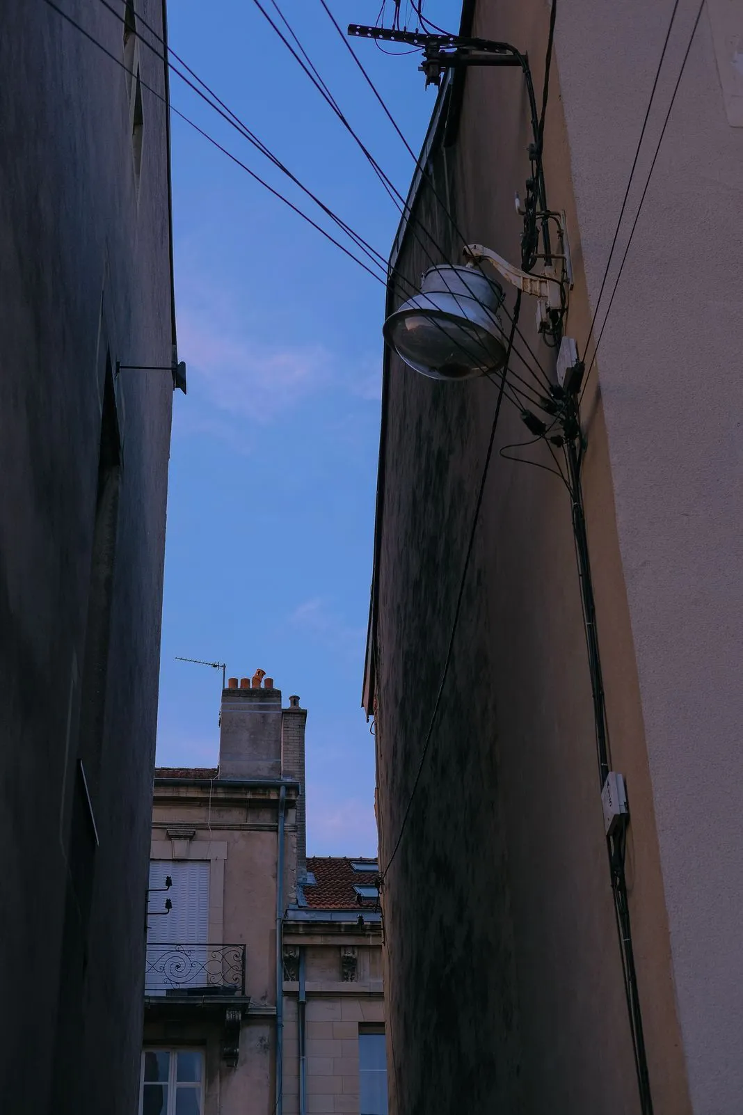 Looking up a narrow alley between buildings at a lamp, wires, and rooftop chimneys