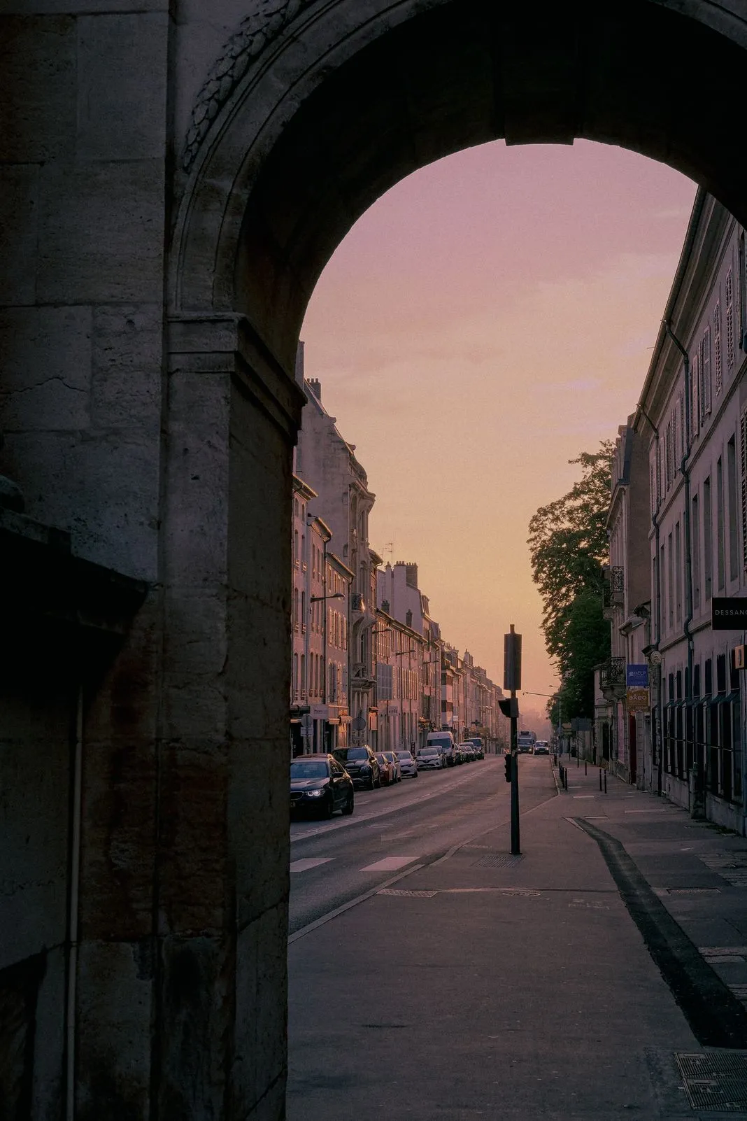 View through a stone archway onto a street lined with buildings under a glowing sunset sky