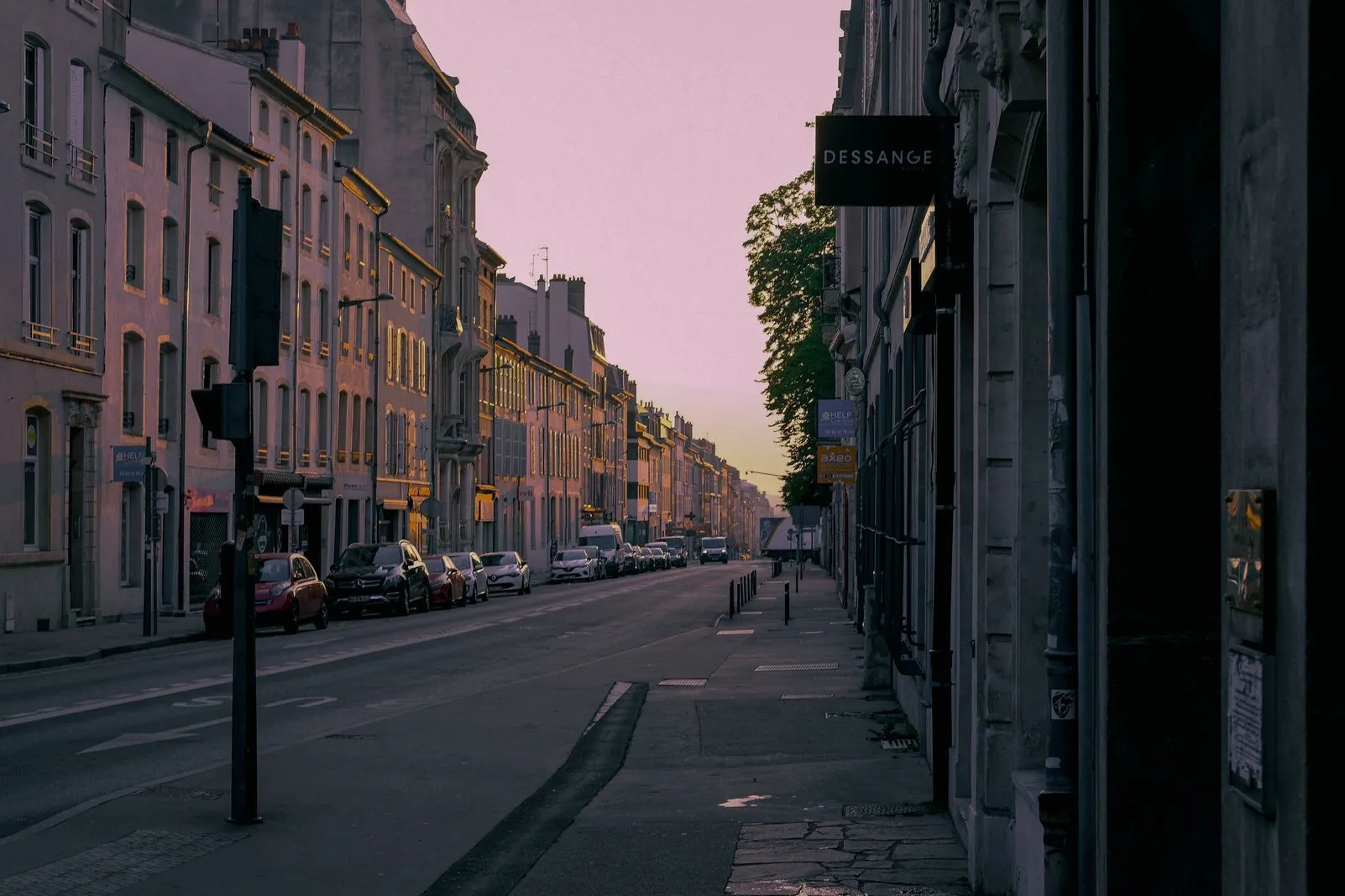 A pink-hued dusk street lined with shops and apartment buildings