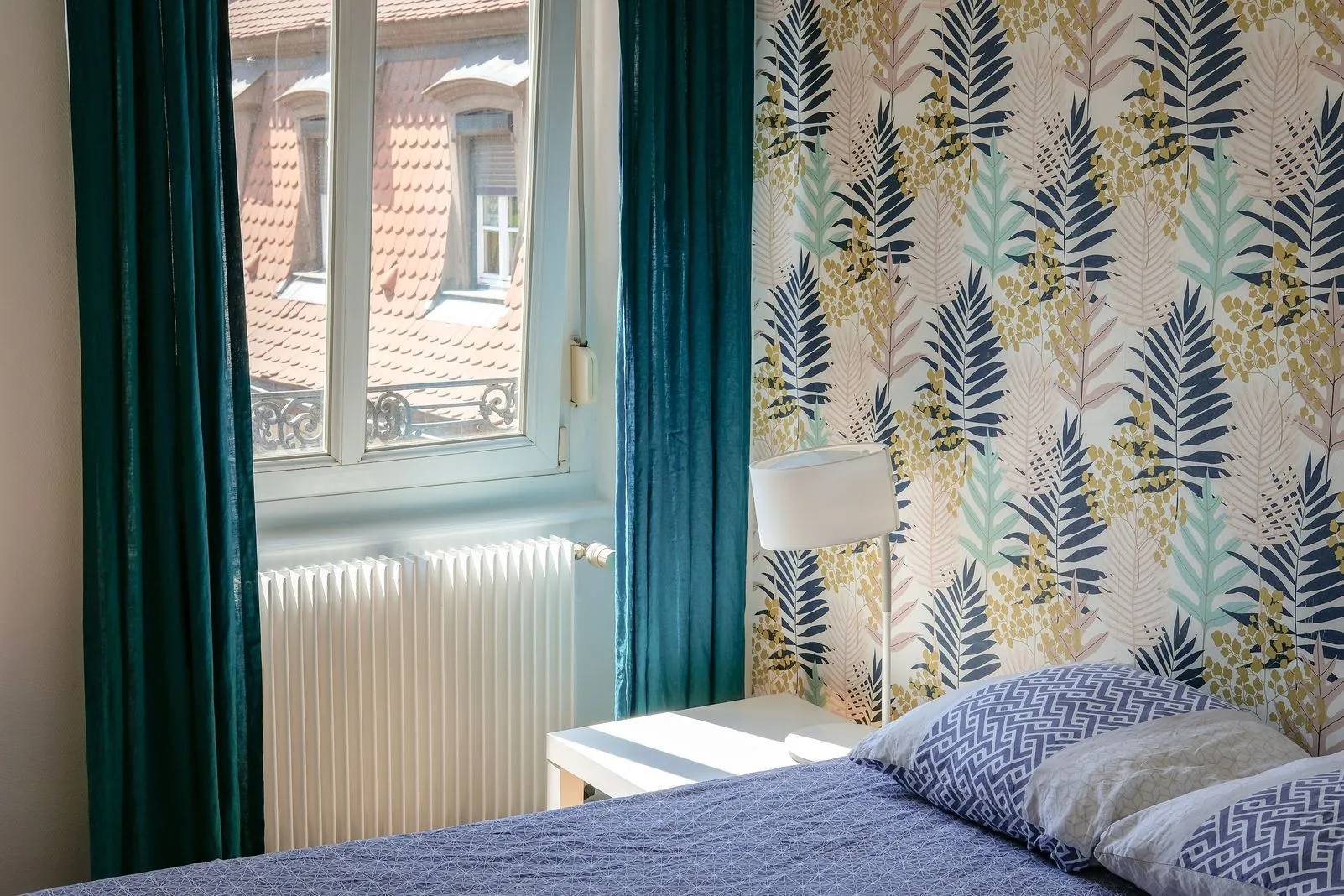 Bedroom with leaf-print wallpaper and a window overlooking terracotta rooftops