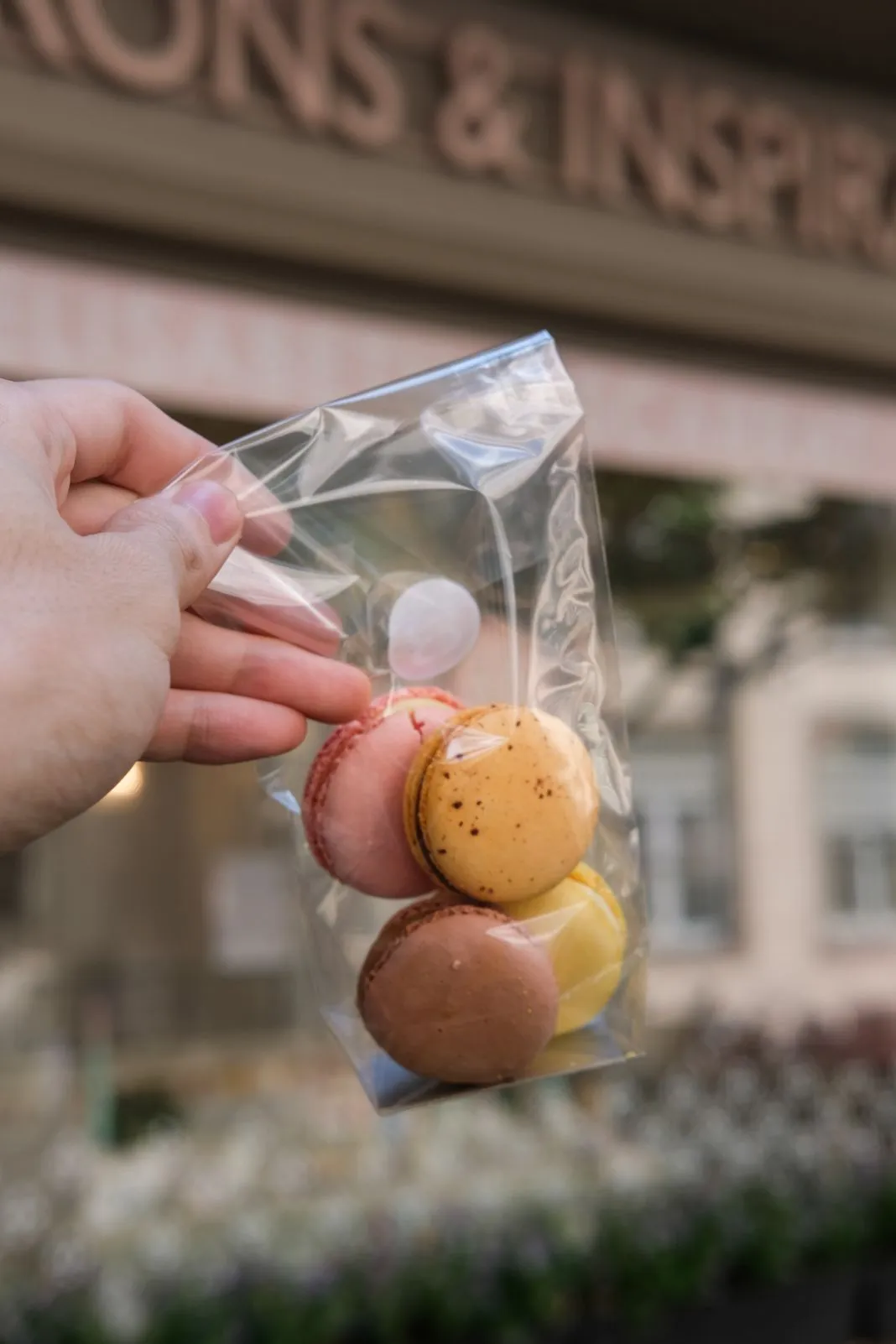 A hand holds a small bag of colorful macarons outside a patisserie