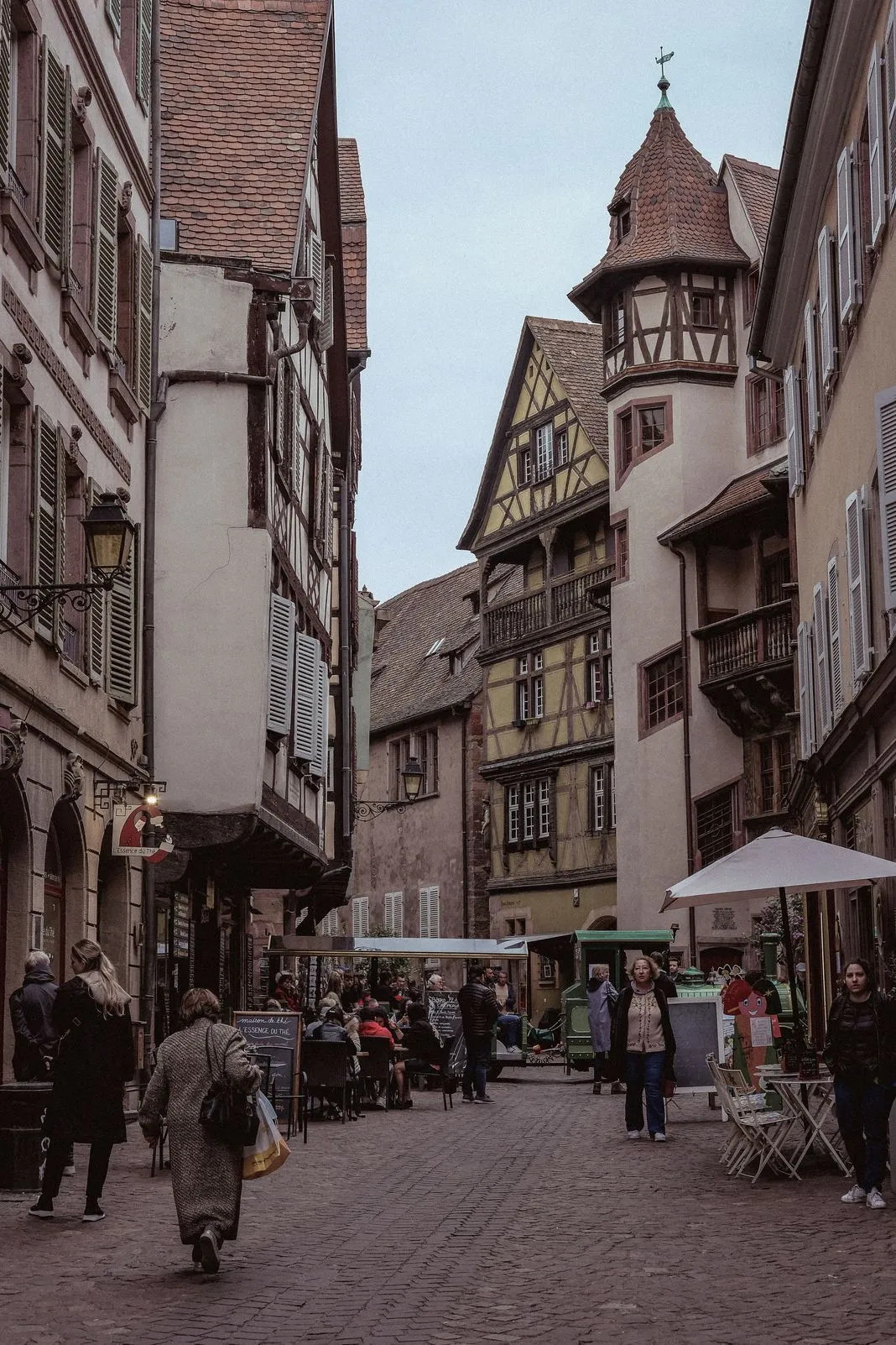 A turreted half-timbered tower rises above a cobbled lane with cafe umbrellas