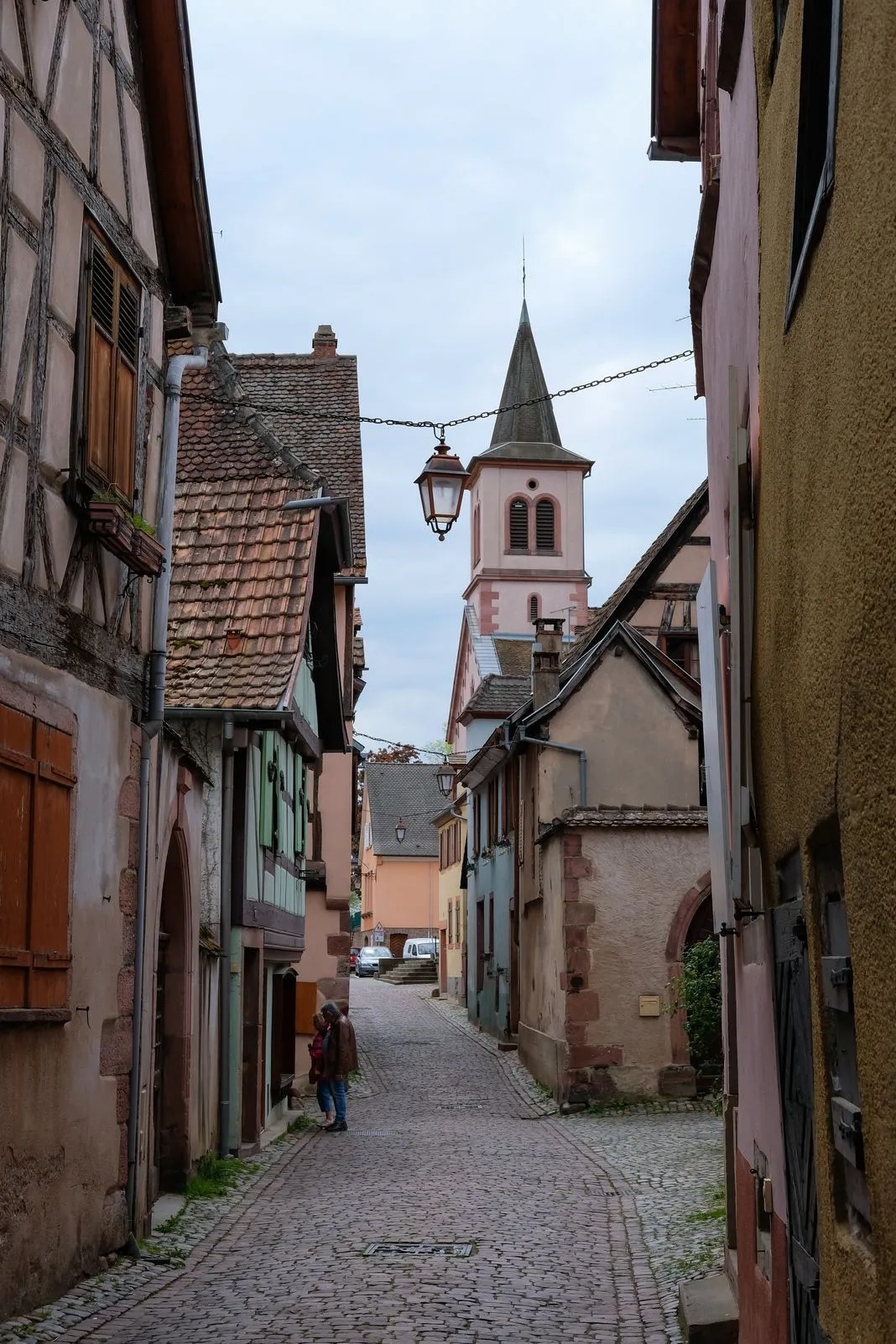 A cobbled lane leads toward a pink church steeple in a half-timbered village