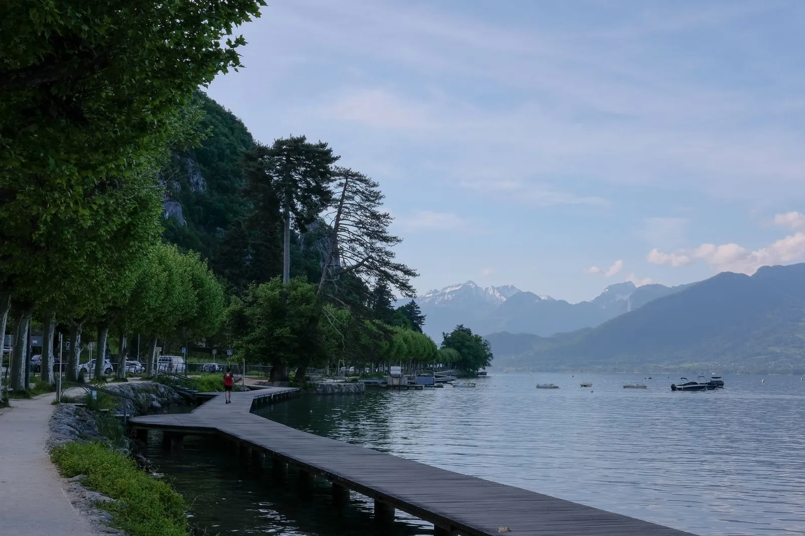 A wooden boardwalk winds along Lake Annecy's shoreline toward snow-capped peaks