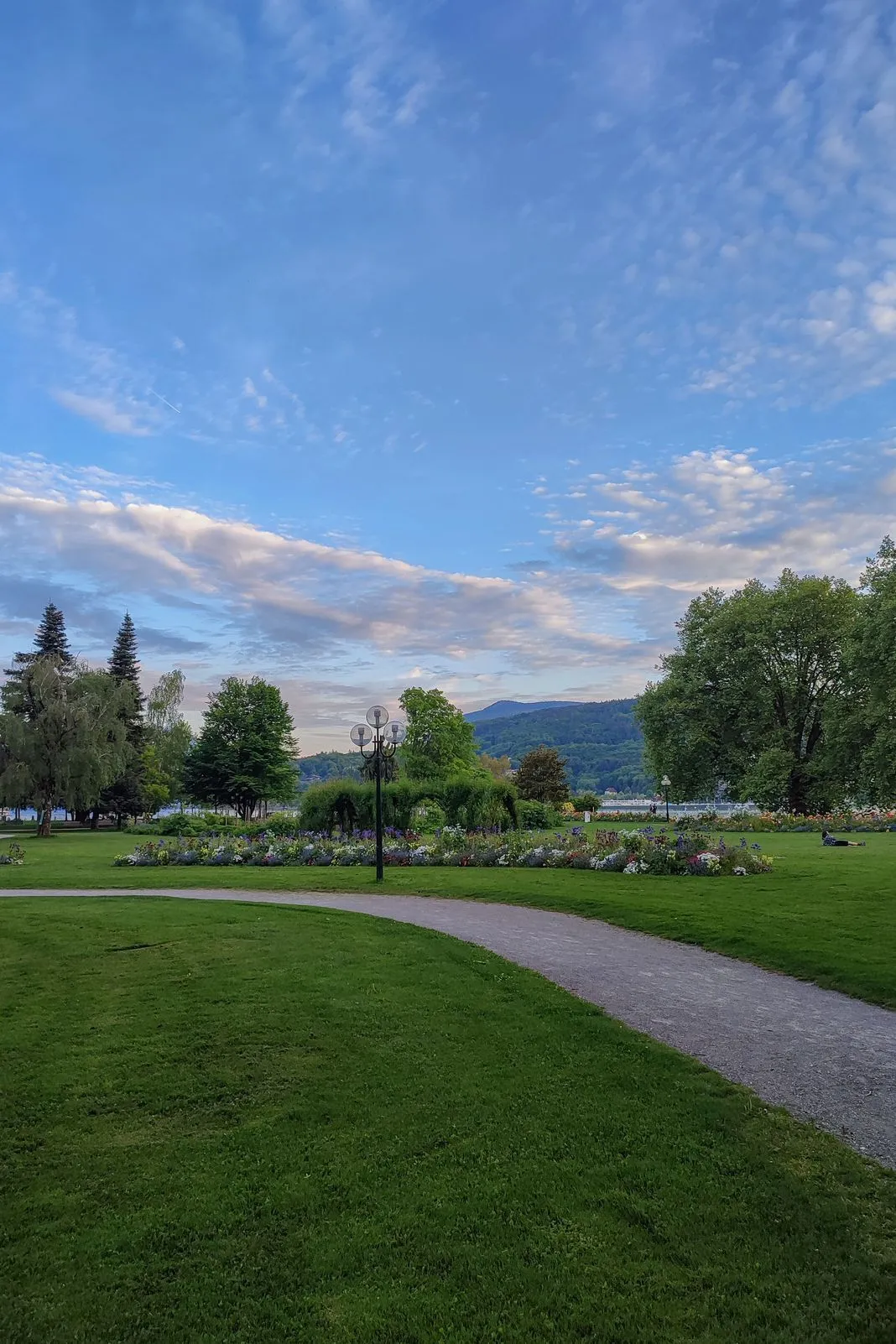 A four-globe lamppost stands beside flower beds in a lakeside park at dusk