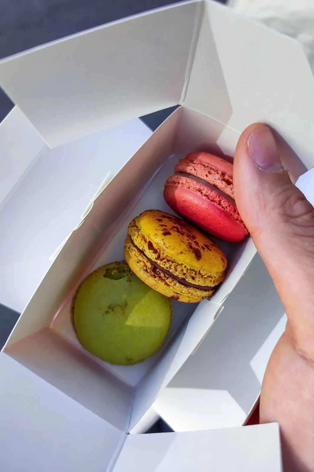 A hand lifts a pink macaron from a box holding pink, yellow, and green ones