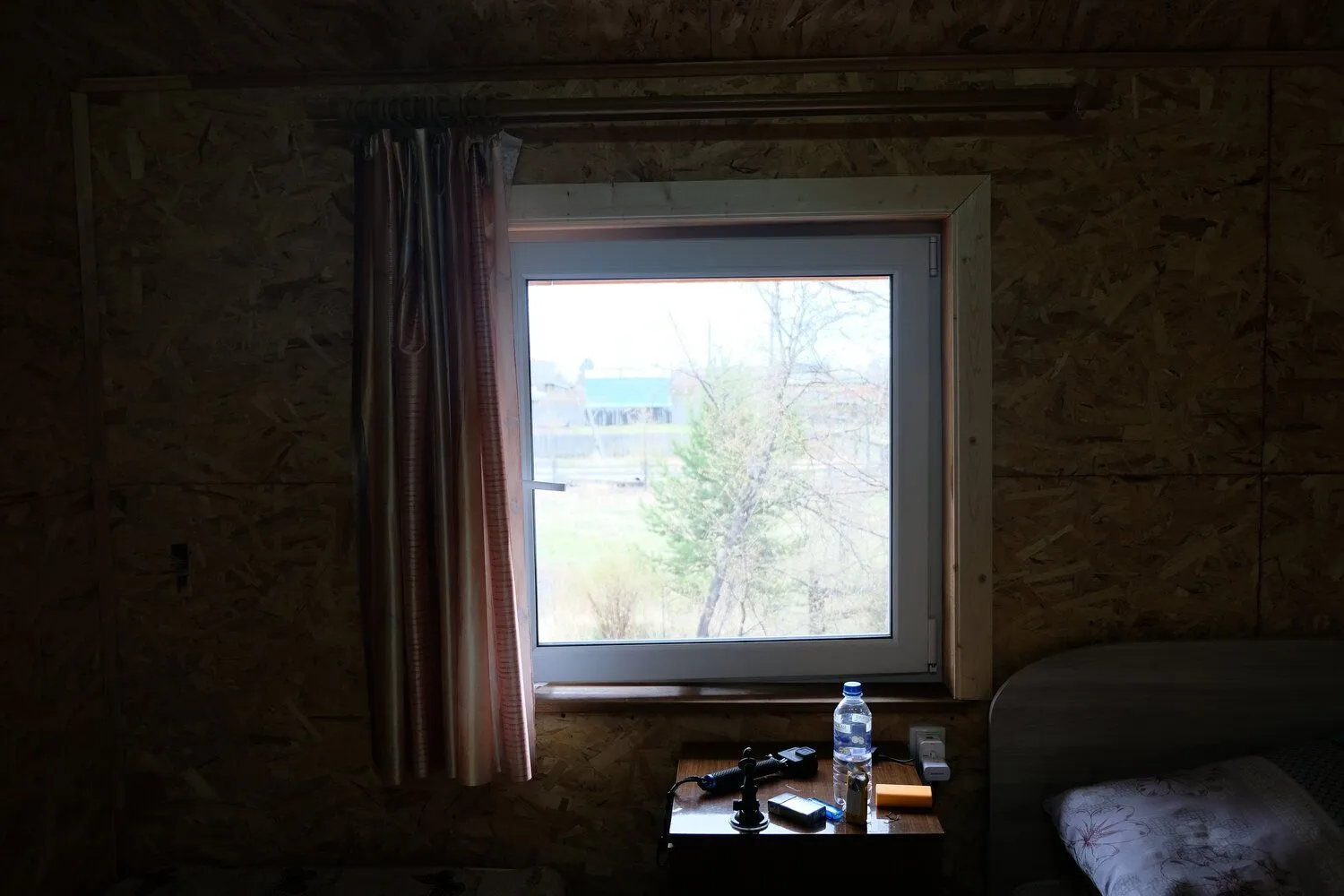 Curtained cabin window looking out over trees, water bottle on the nightstand