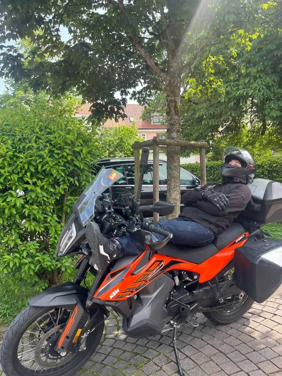 Rider leaning back on an orange KTM adventure bike with side cases parked under a tree