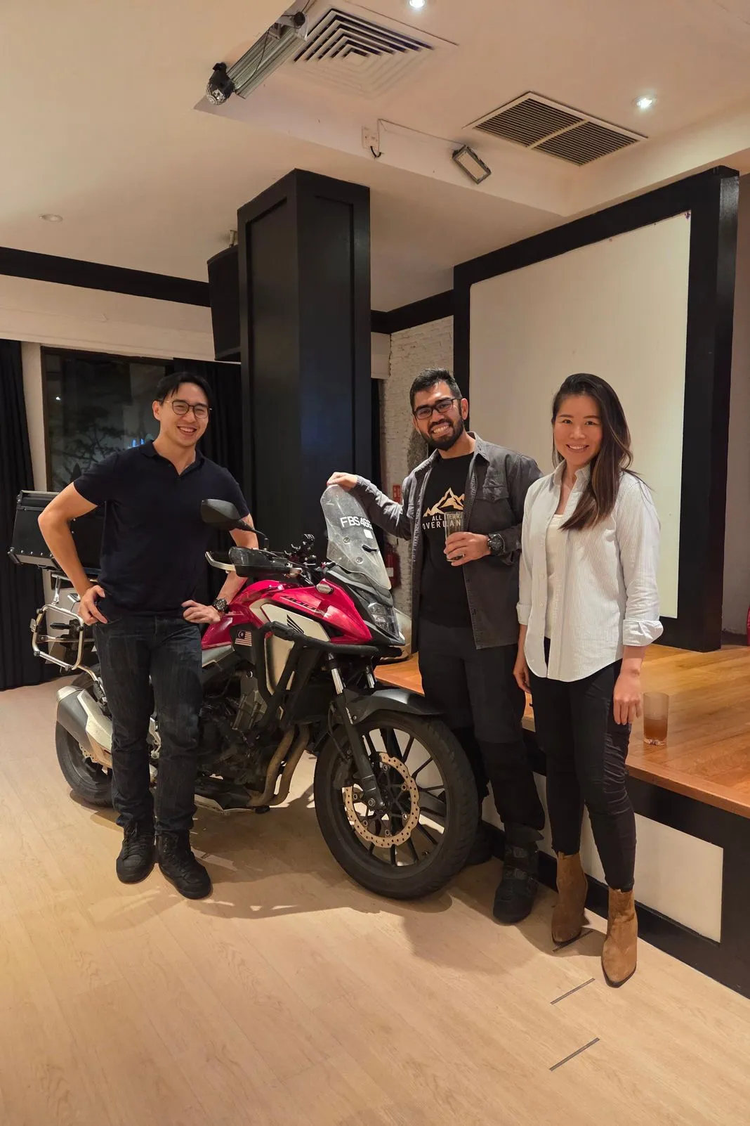 Three people posing with a red Honda adventure bike indoors at an evening meetup