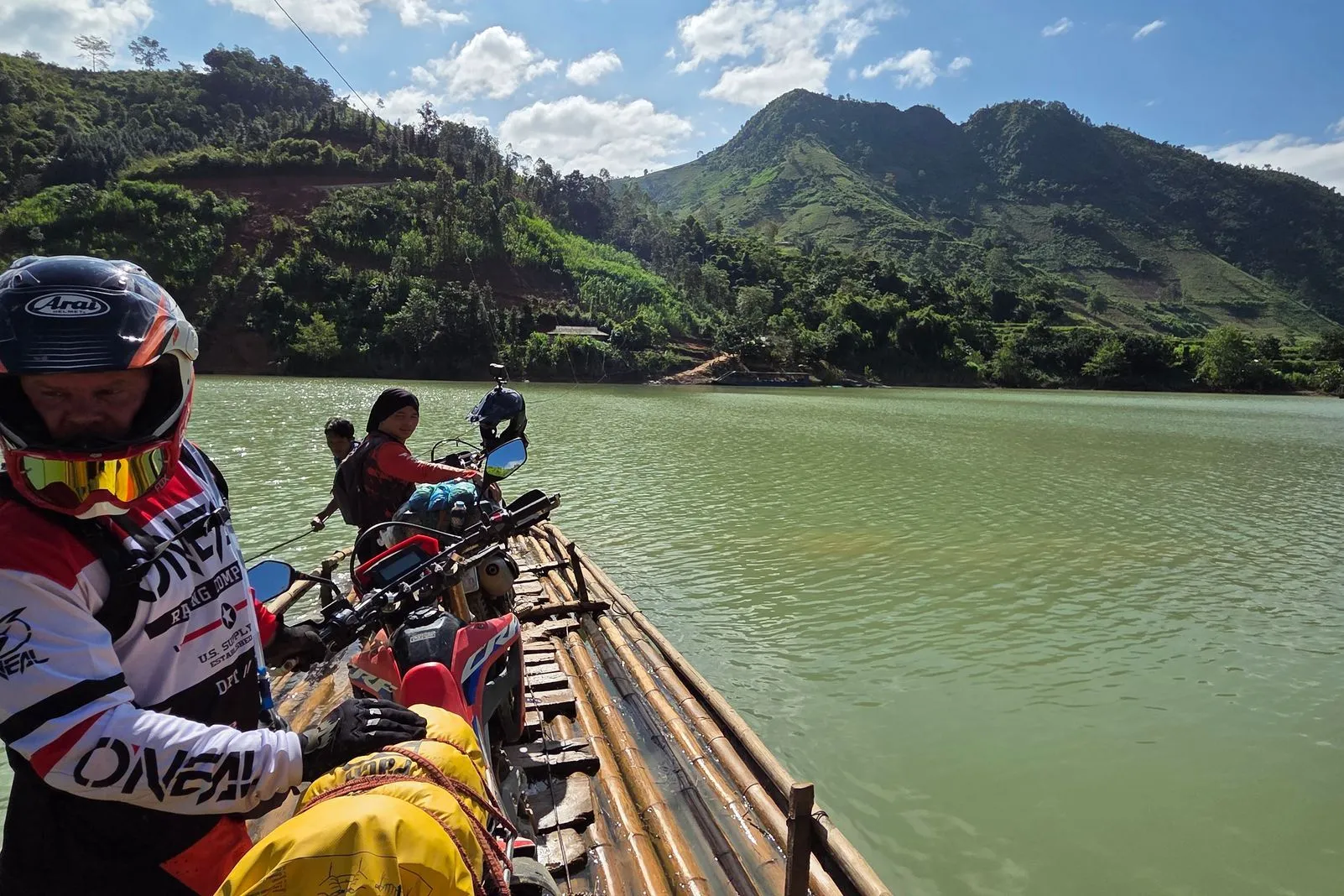 Dirt bikes loaded on a bamboo raft crossing a green river below jungle-covered mountains