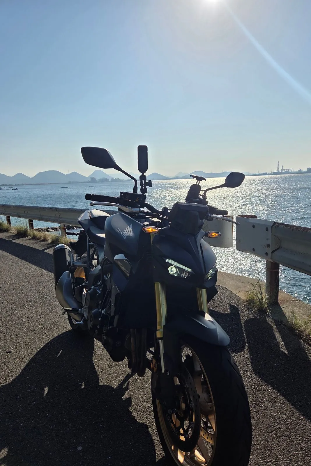 Black Honda naked bike parked at a guardrail overlooking a sunlit bay and distant hills