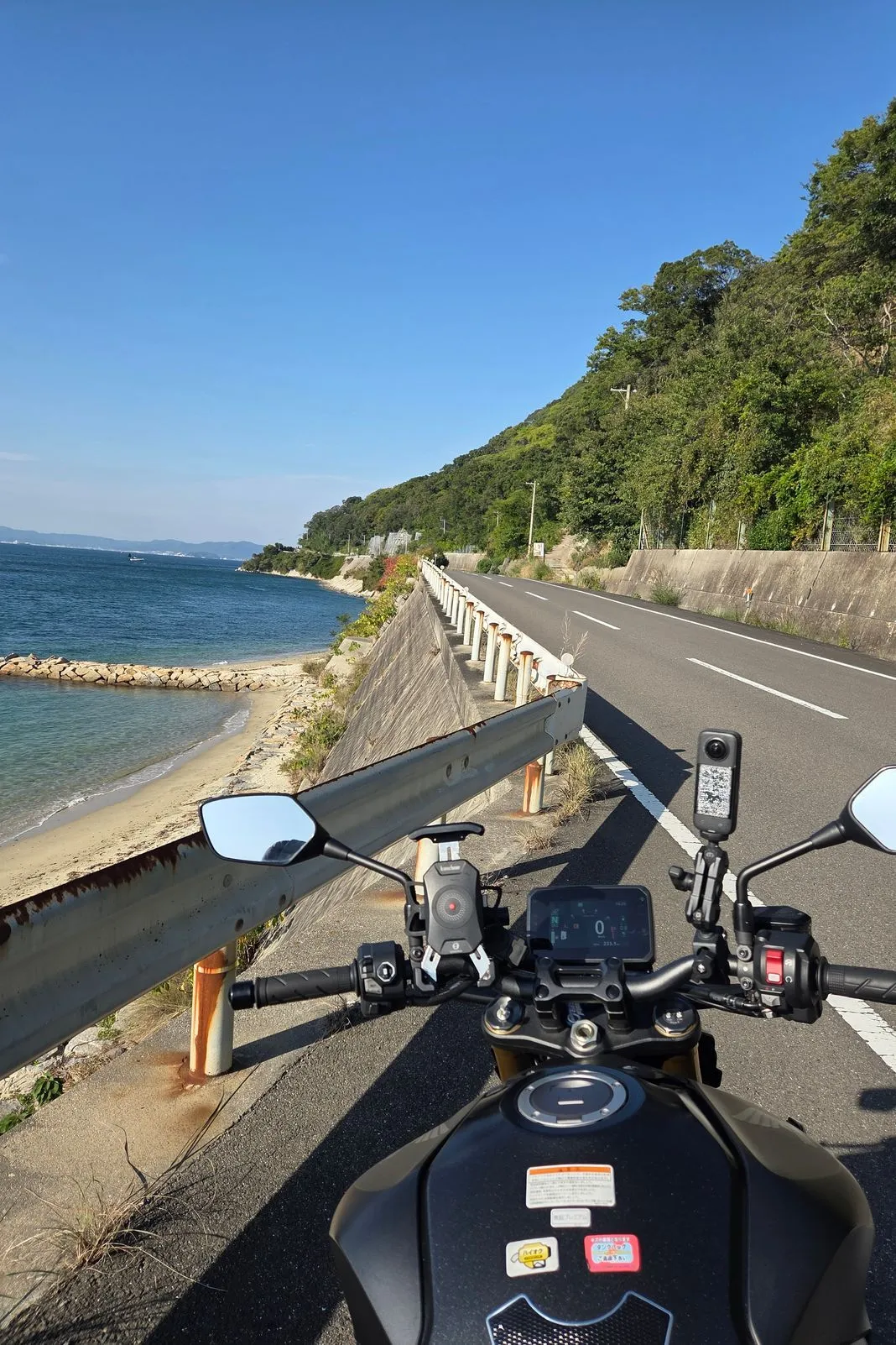 Handlebar-view riding a coastal road with a guardrail, sandy cove, and forested hillside