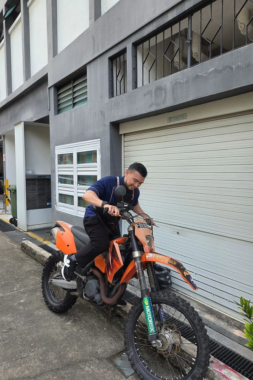 Smiling rider straddling an orange KTM enduro bike in a back lane between buildings