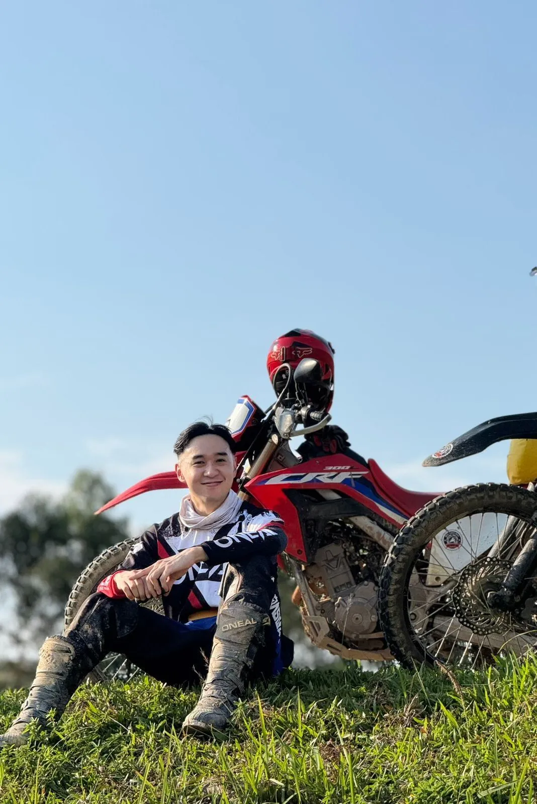 Rider in muddy gear sitting on a grassy hill beside a red Honda CRF300L dirt bike