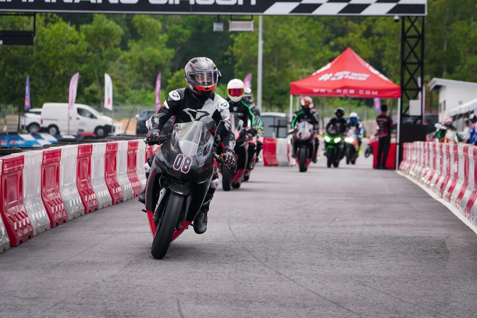 Pack of riders exiting pit lane under a checkered start-finish gantry at a race circuit
