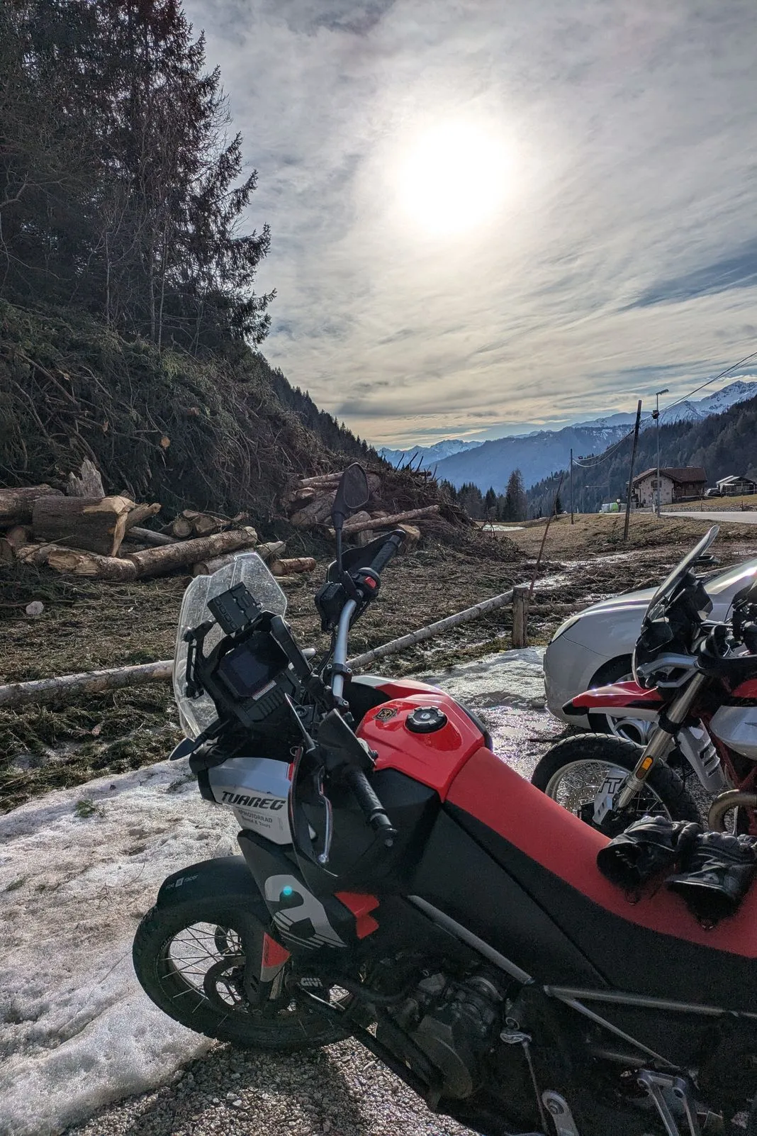 Aprilia Tuareg parked beside a stacked log pile with snow-capped peaks lit by low sun