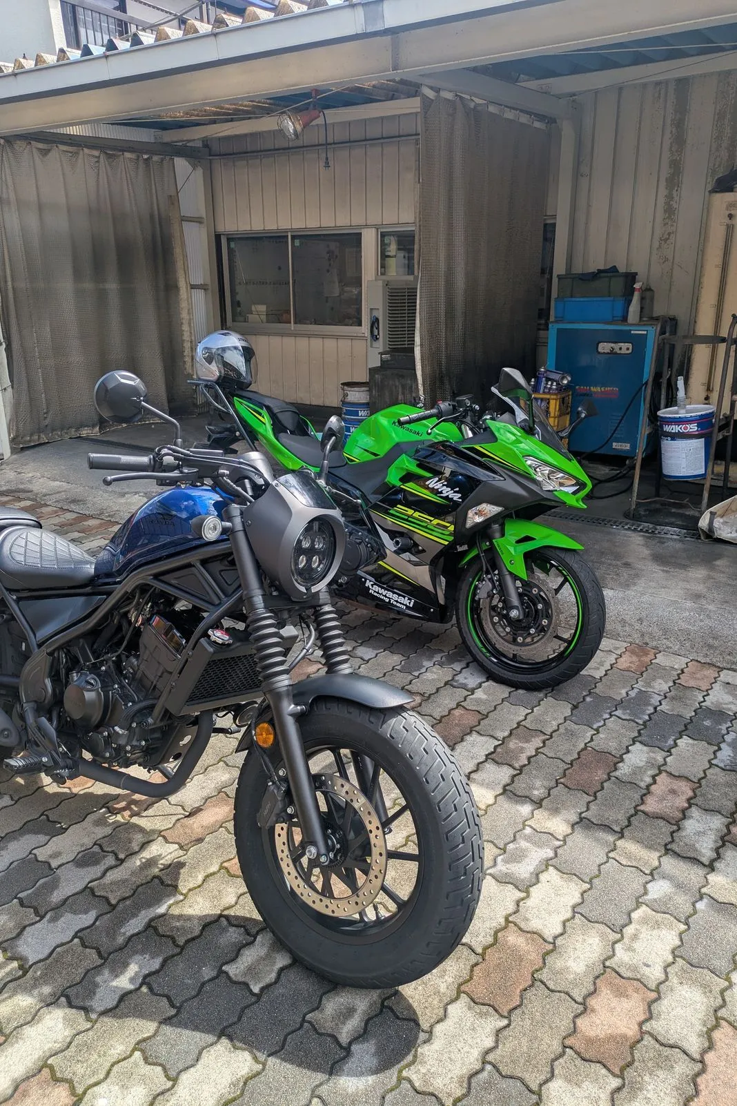 Green Kawasaki Ninja 250 parked beside a blue cafe-style motorcycle under a carport