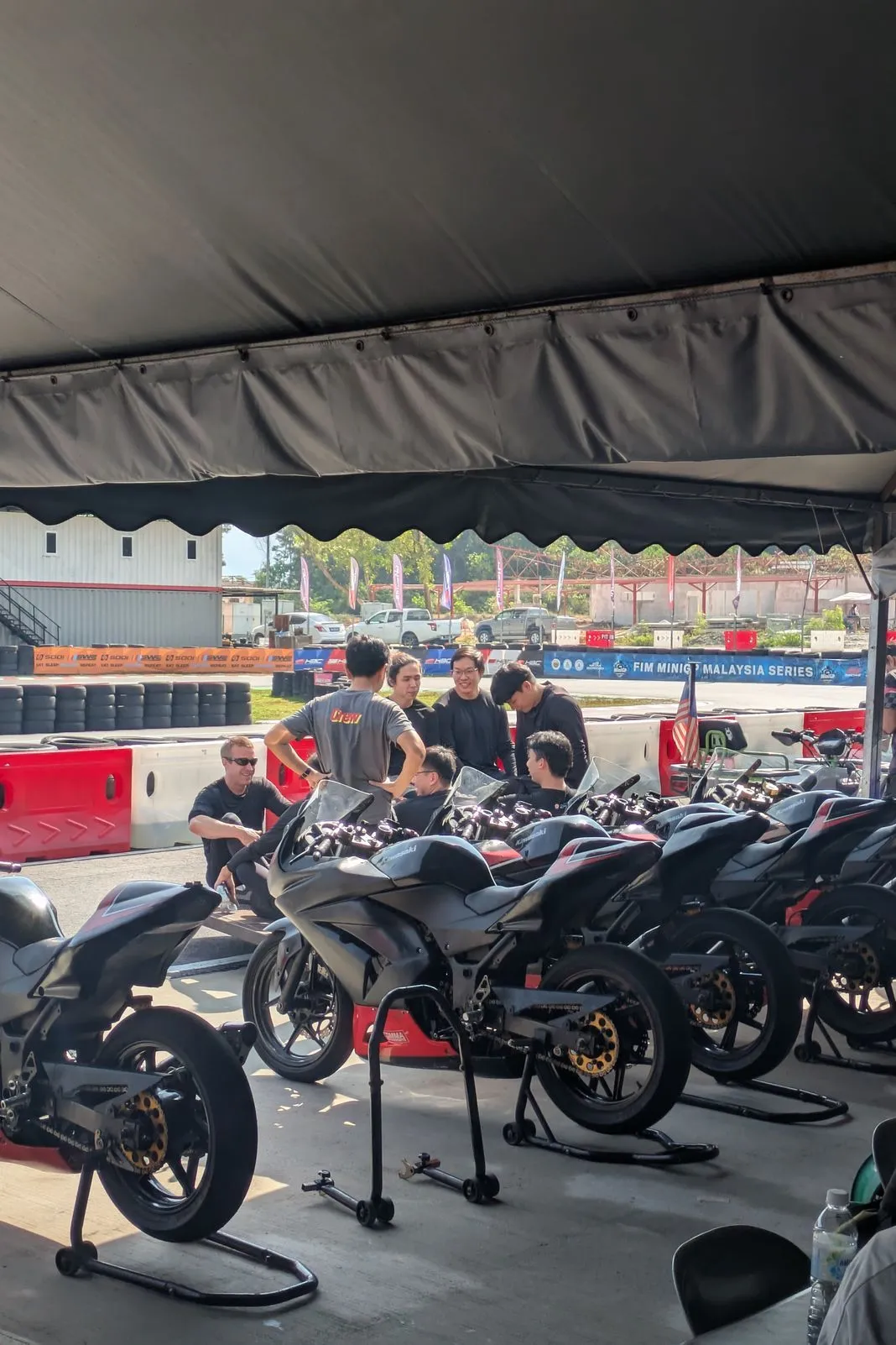 Row of race-prepped sportbikes on paddock stands under a tent as riders chat at a track day