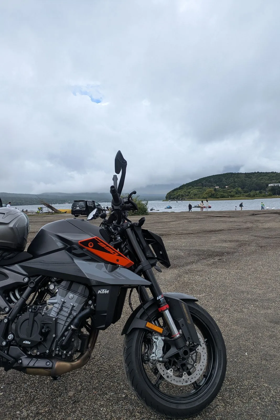 Black and orange KTM parked on a lakeside gravel lot with hazy hills across the water