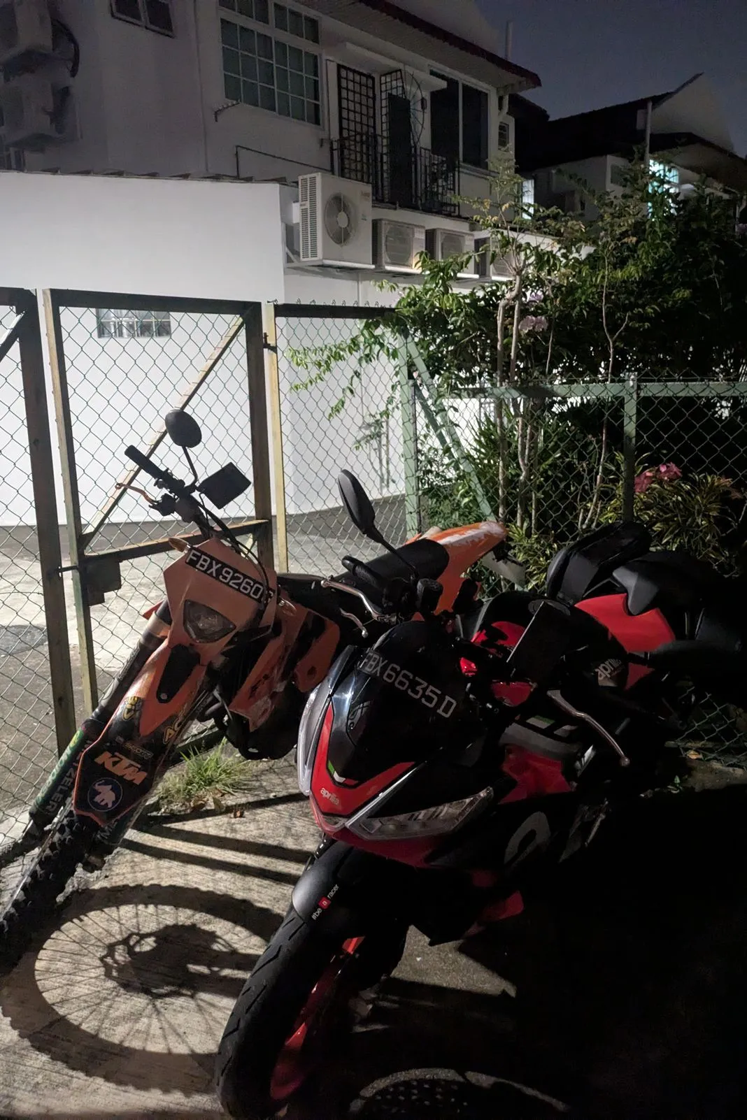 Orange KTM dirt bike and a black-and-red Aprilia parked together at night behind a chain-link gate