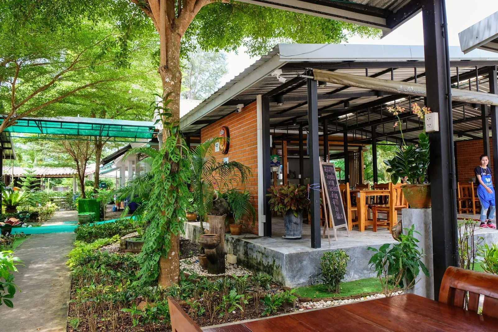 Outdoor restaurant terrace with a brick building, tropical plants, and wooden tables