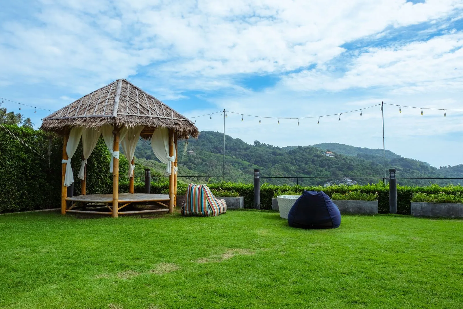 Thatched-roof gazebo with white curtains on a lawn, string lights strung across the yard