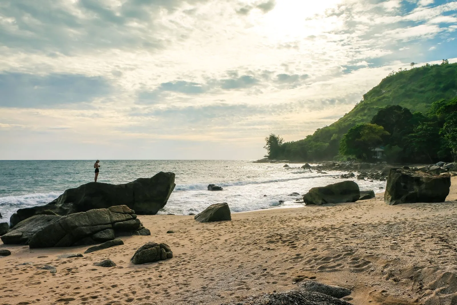 Person standing on a large rock at the beach as the sun sets behind clouds