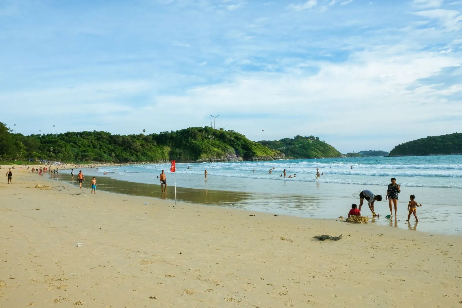 Wide sandy beach with swimmers, sunbathers, and a red flag, a headland in the distance