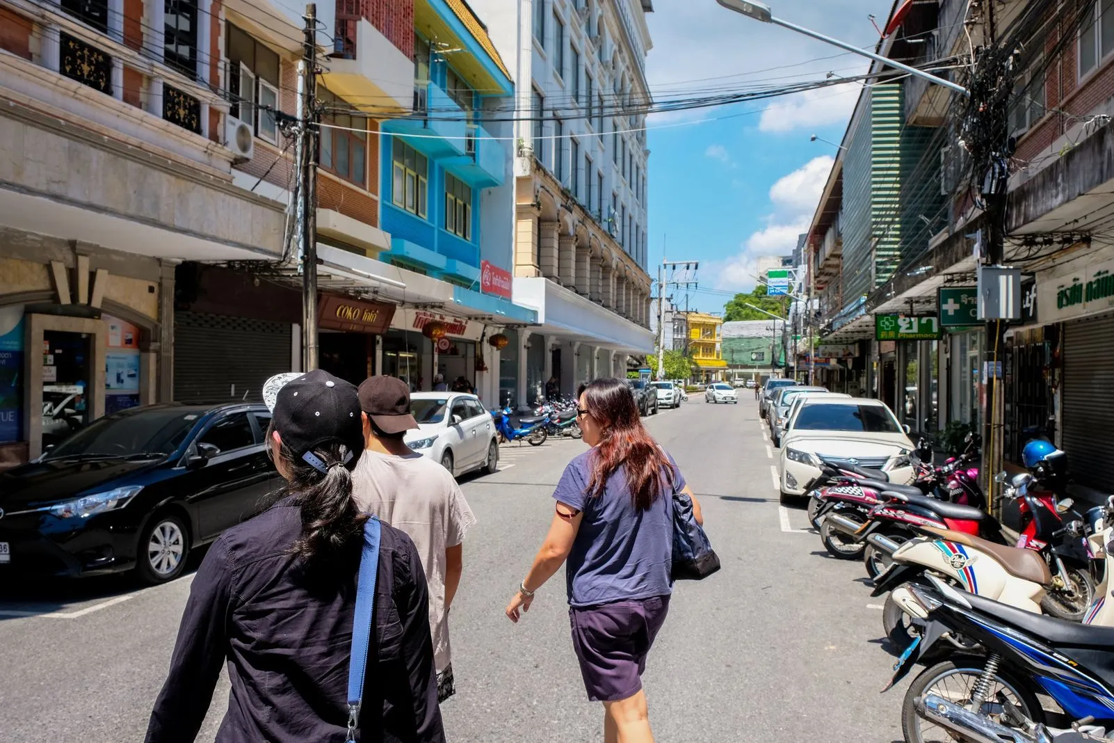 People walking down a street lined with parked cars and motorbikes