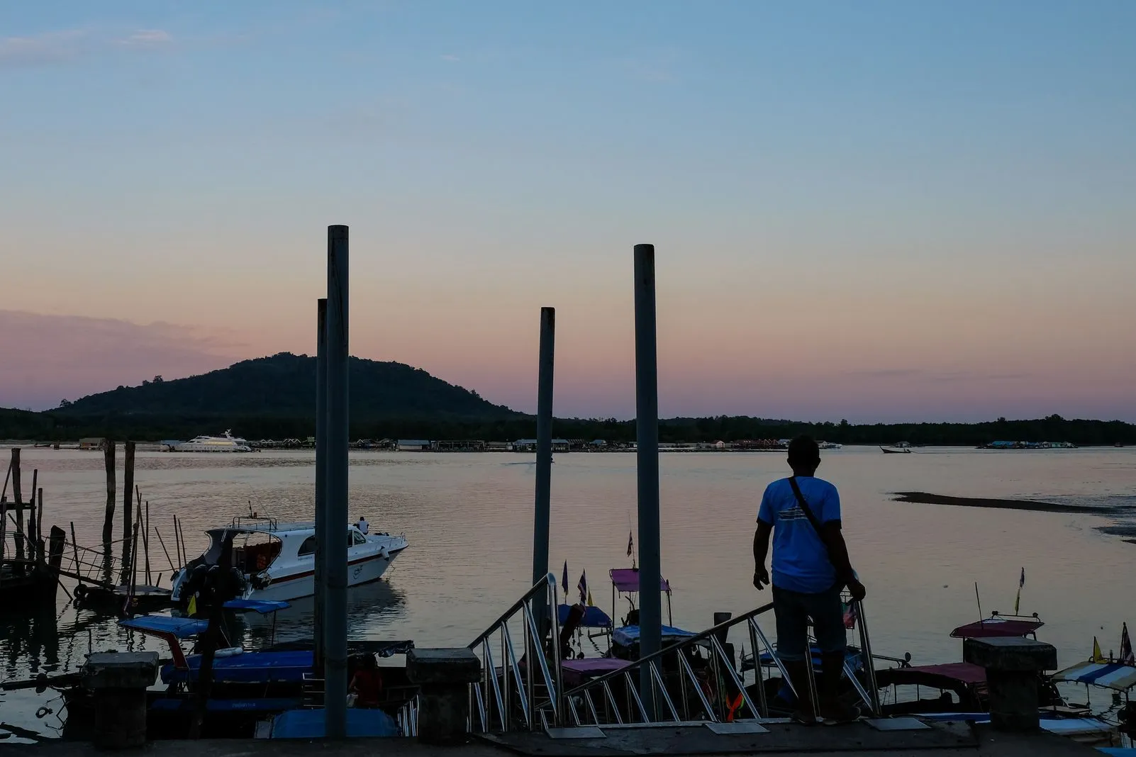 Silhouette of a person standing on a jetty at dusk among mooring poles and small boats