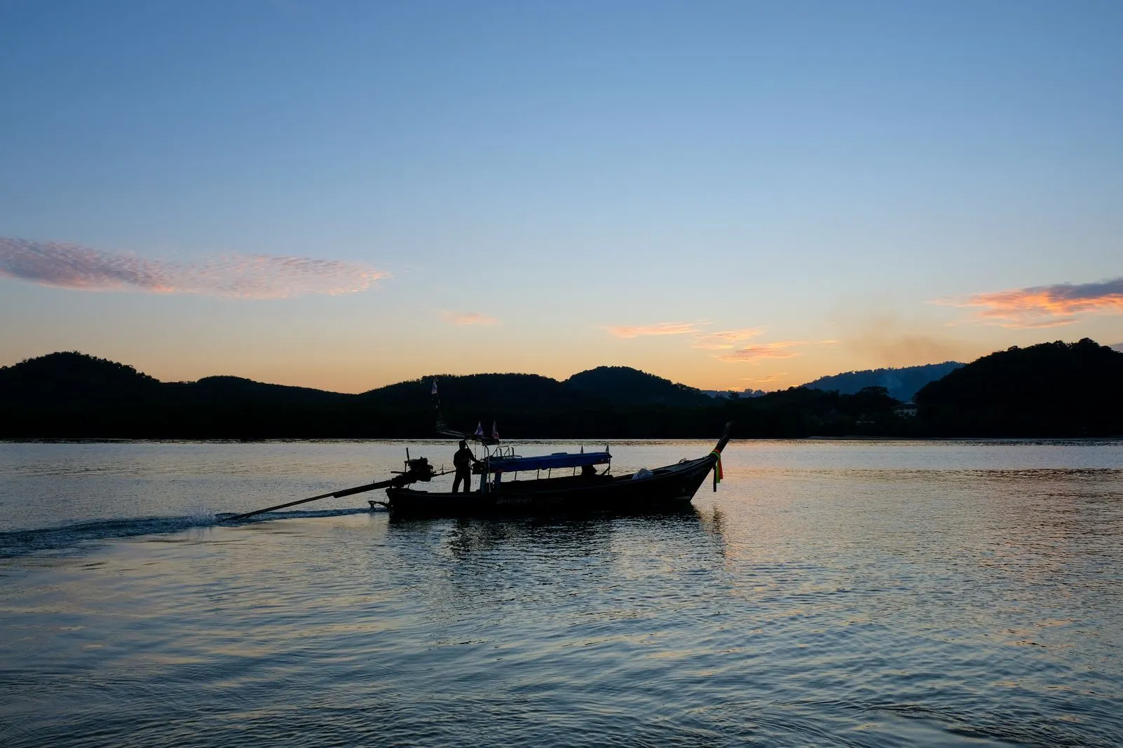 Silhouette of a longtail boat crossing calm water at sunset with hills behind