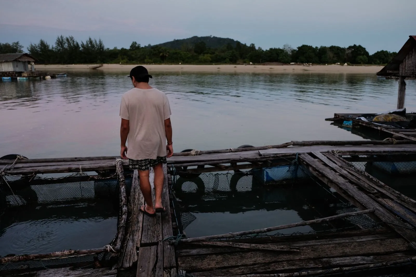 Person walking along a floating wooden dock past fish pens, a beach beyond