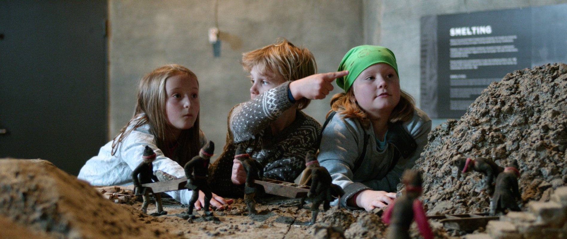 Tre barn utforsker en detaljert modellutstilling på museum. I forgrunnen vises små figurer og landskap som illustrerer arbeid ved et historisk smelteverk, mens barna studerer modellen på nært hold.