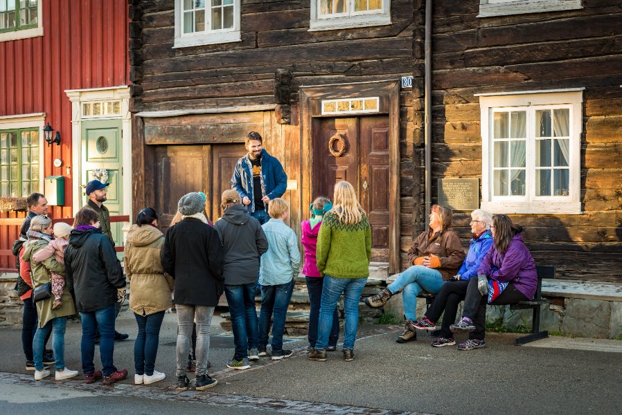 En gruppe besøkende står samlet utenfor gamle trehus i et historisk gatemiljø og lytter til en omvisning. Noen sitter på en benk ved husveggen, mens resten står rundt guiden i gata.