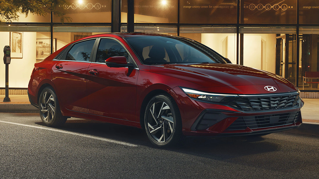 Vue 3/4 avant et latérale du Hyundai Elantra 2025, garé devant un batîment luxueux avec phares allumées