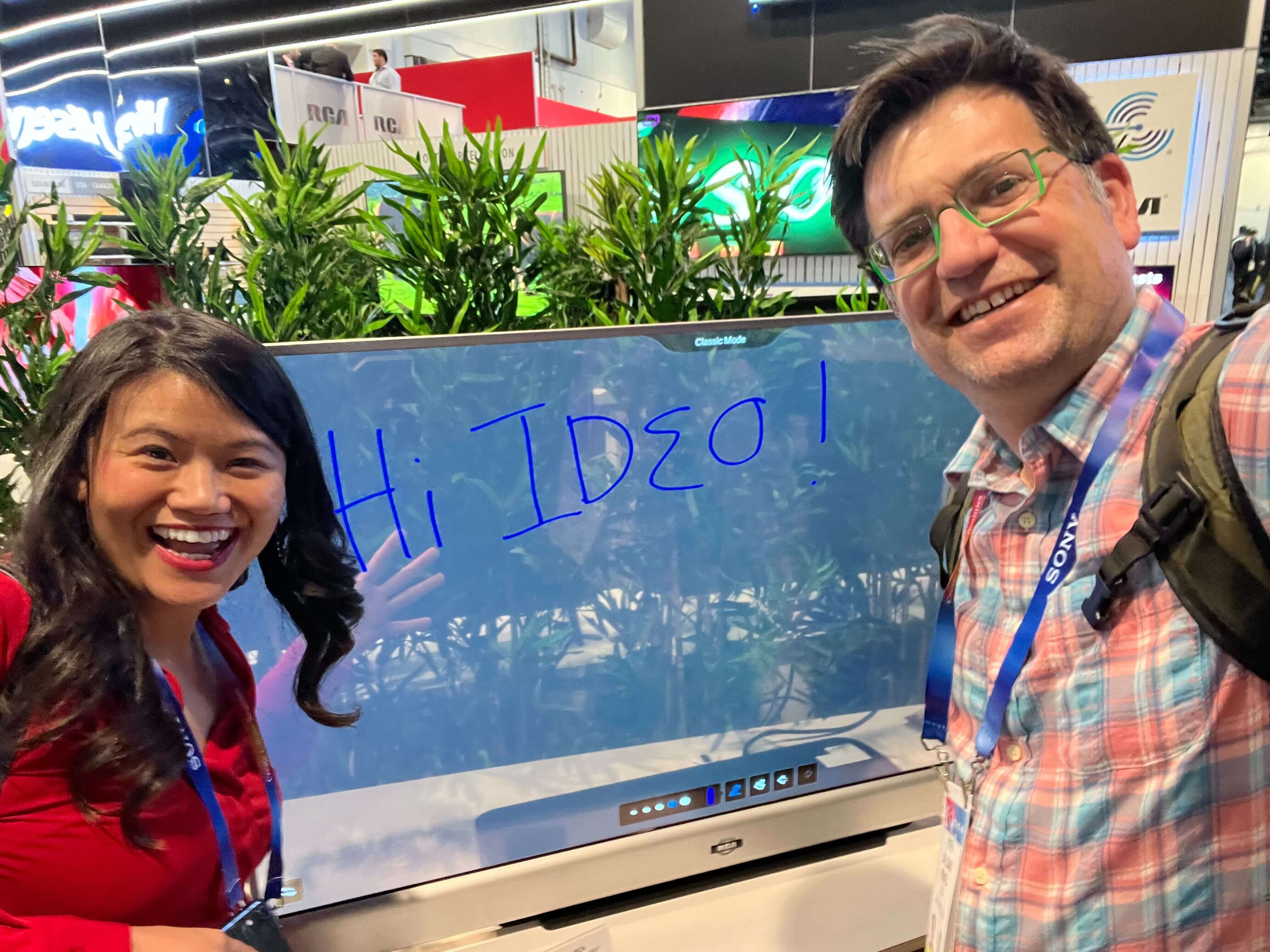 Smiling individuals at CES booth: Two people, one woman and one man, pose smiling for a photo next to an interactive display screen that reads "Hi IDEO!" in blue text.