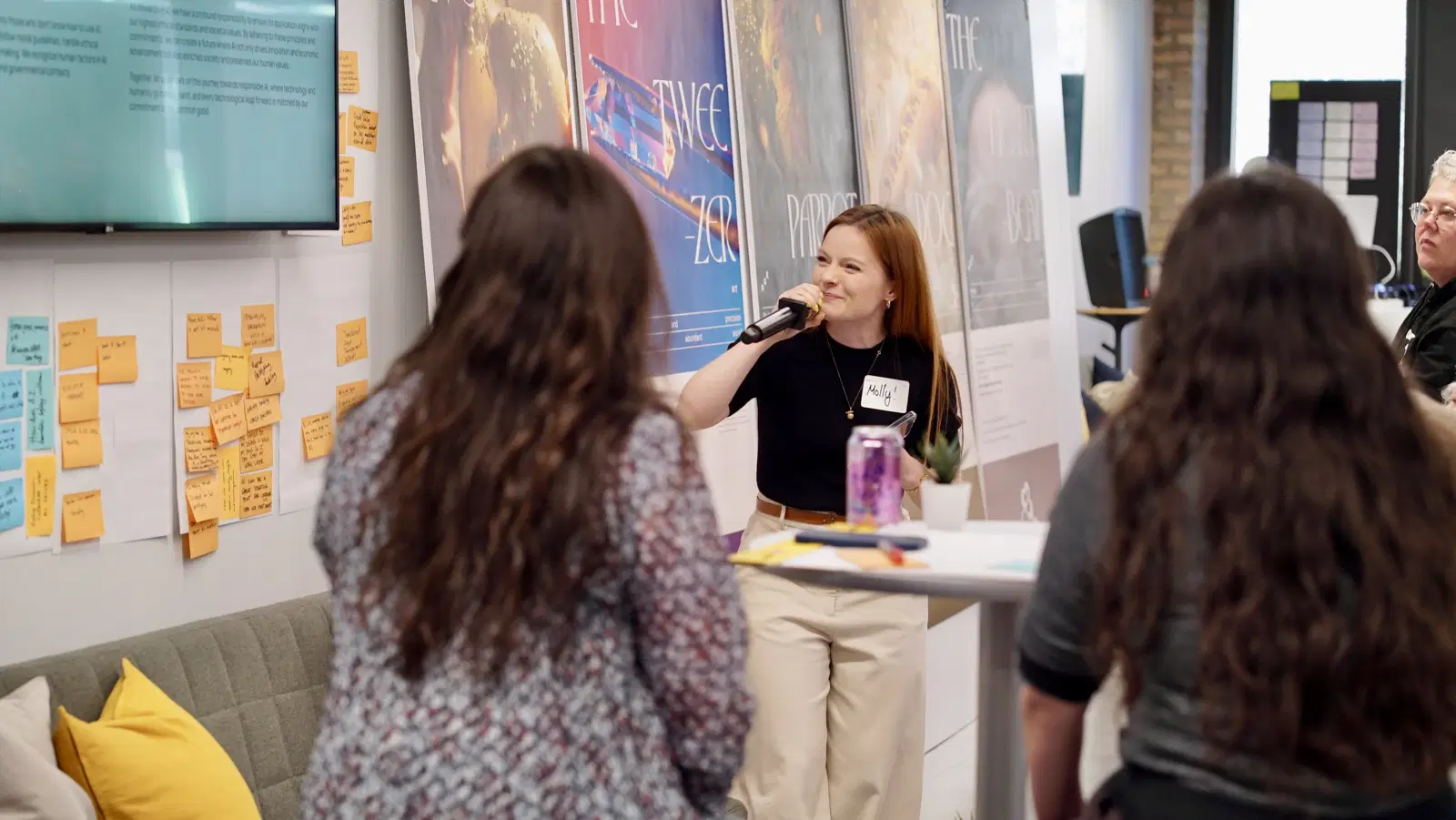 A person speaking in to a microphone at an event, posters are in the background and attendees are in the foreground