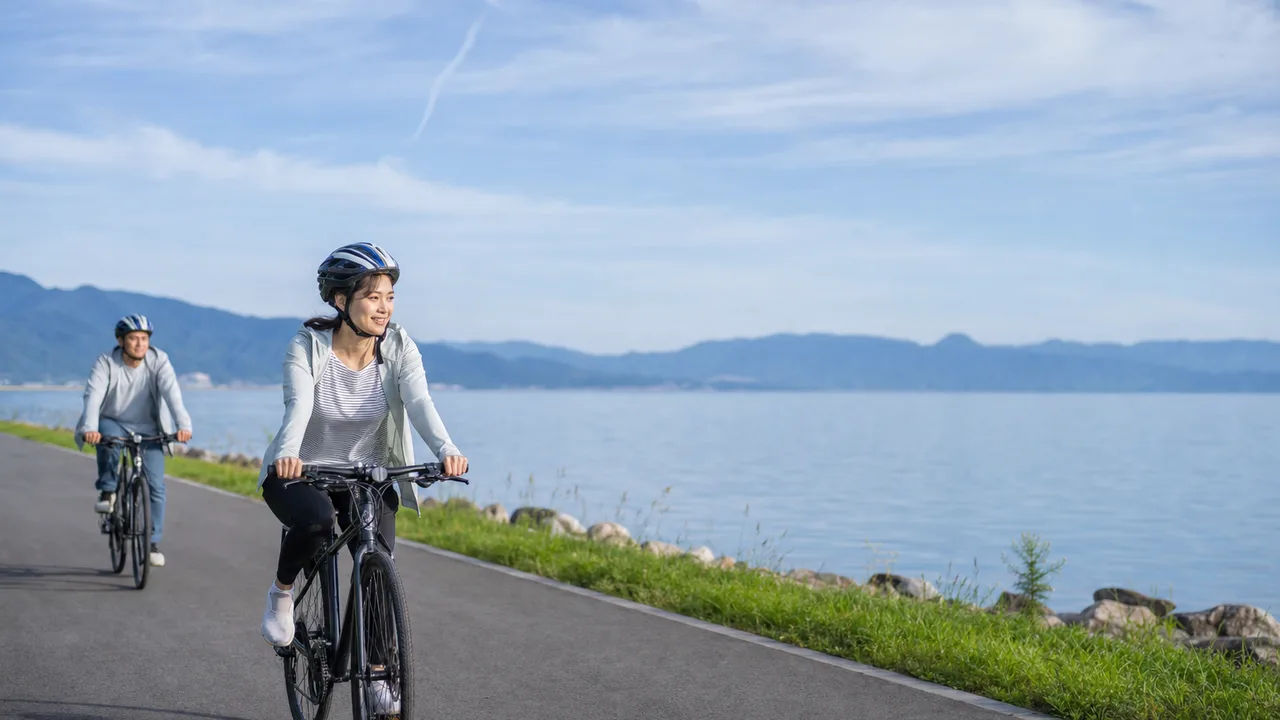 滋賀の琵琶湖畔を自転車で巡る旅行者（横長・コピースペース）。滋賀旅行、琵琶湖、サイクリング、自転車旅行の記事や案内に使える商用素材です。