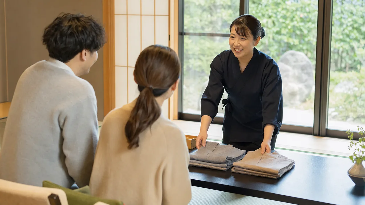 旅館で迎える日本人スタッフと旅行者（横長・コピースペース）。旅館、温泉旅行、おもてなし、国内旅行の記事向け素材です。