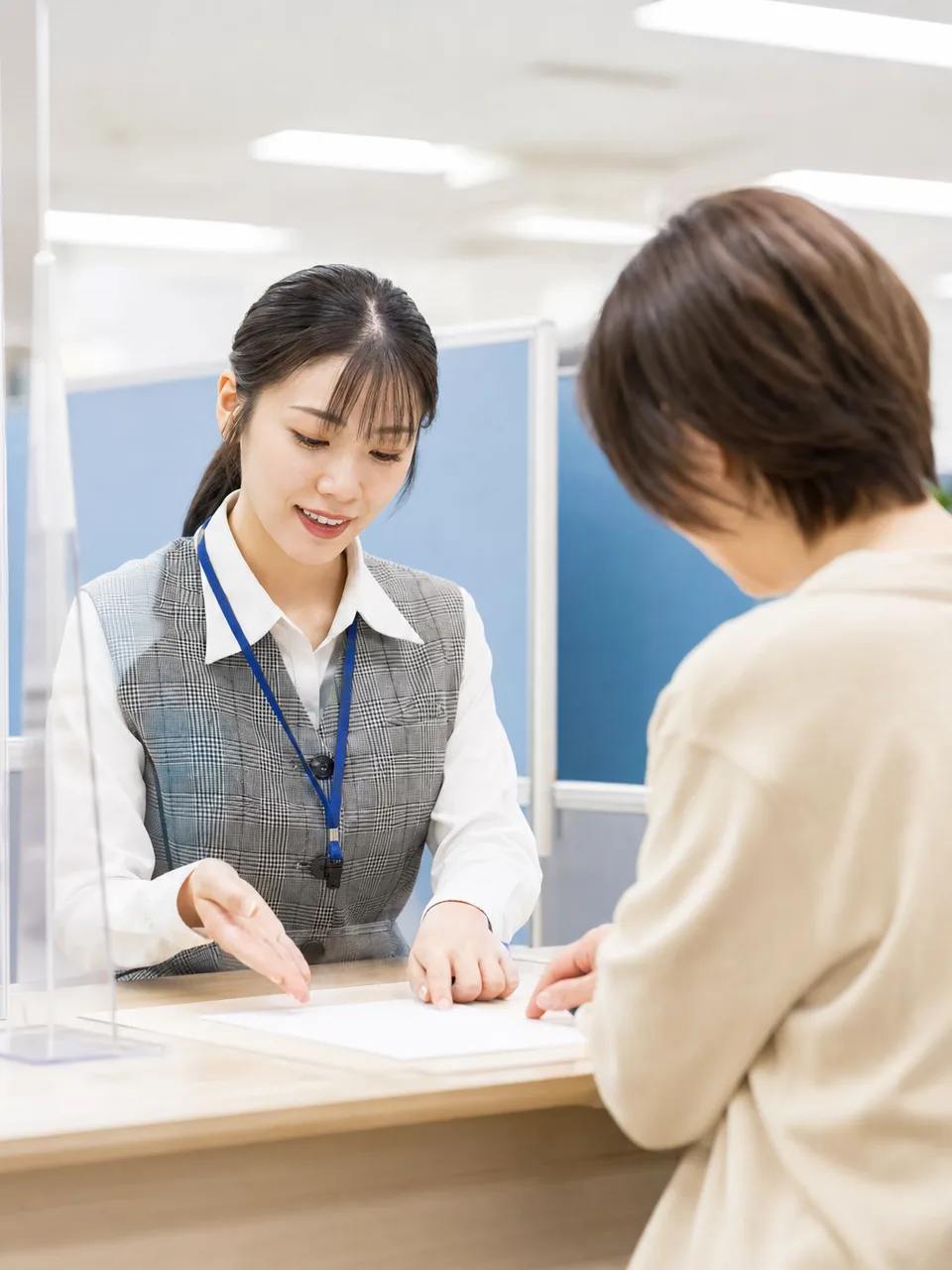 市役所窓口で案内する日本人公務員（自然な縦位置写真）。公務員、市役所、行政サービス、窓口対応の記事向け素材です。