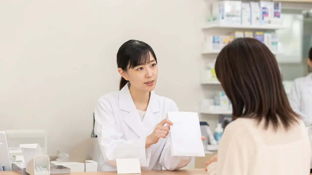 調剤薬局で説明する日本人薬剤師（横長・コピースペース）。調剤薬局、薬剤師、服薬説明、地域医療の記事向け素材です。